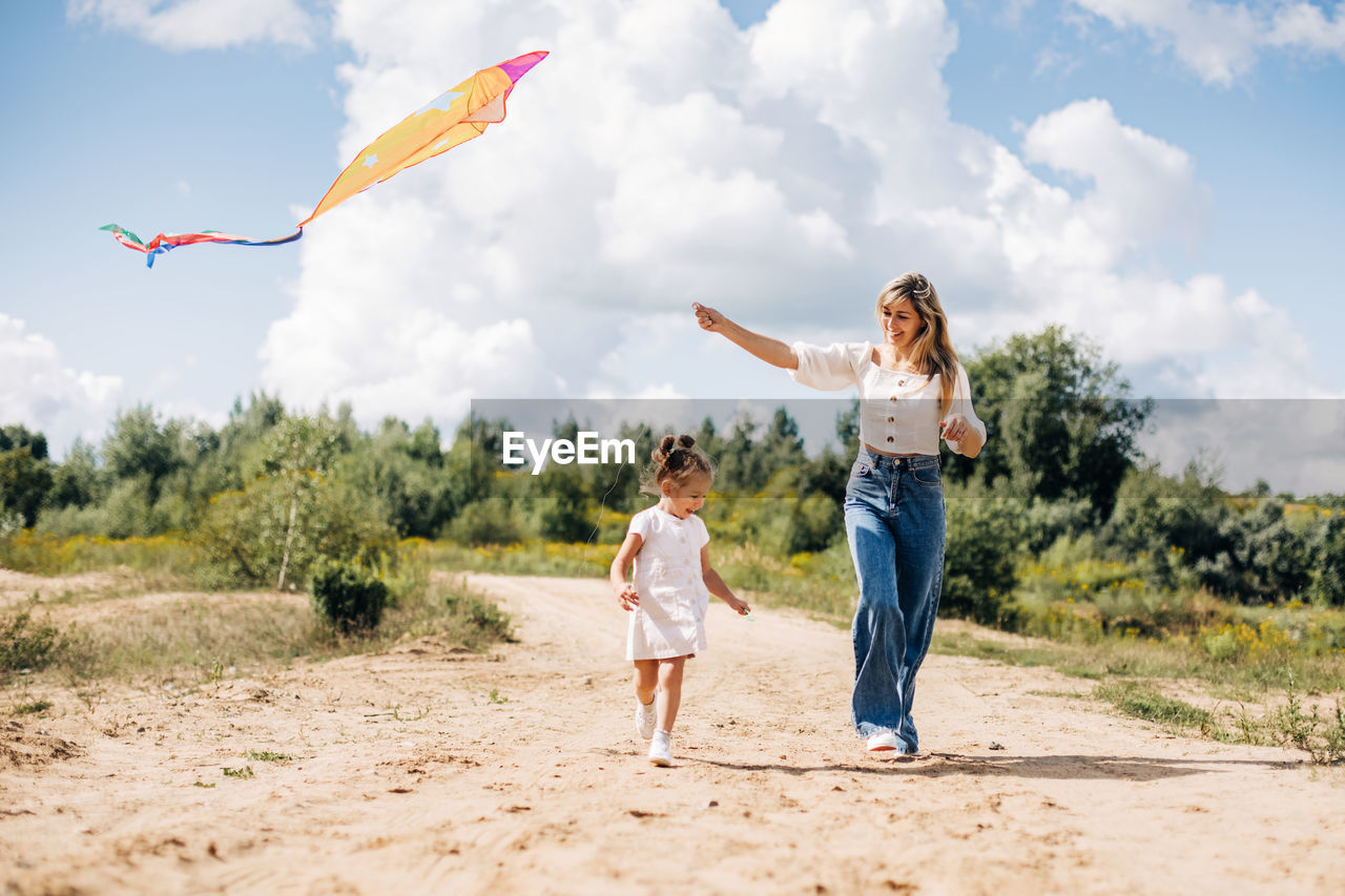 A laughing little girl and her mother run along the road to the field and fly a kite.