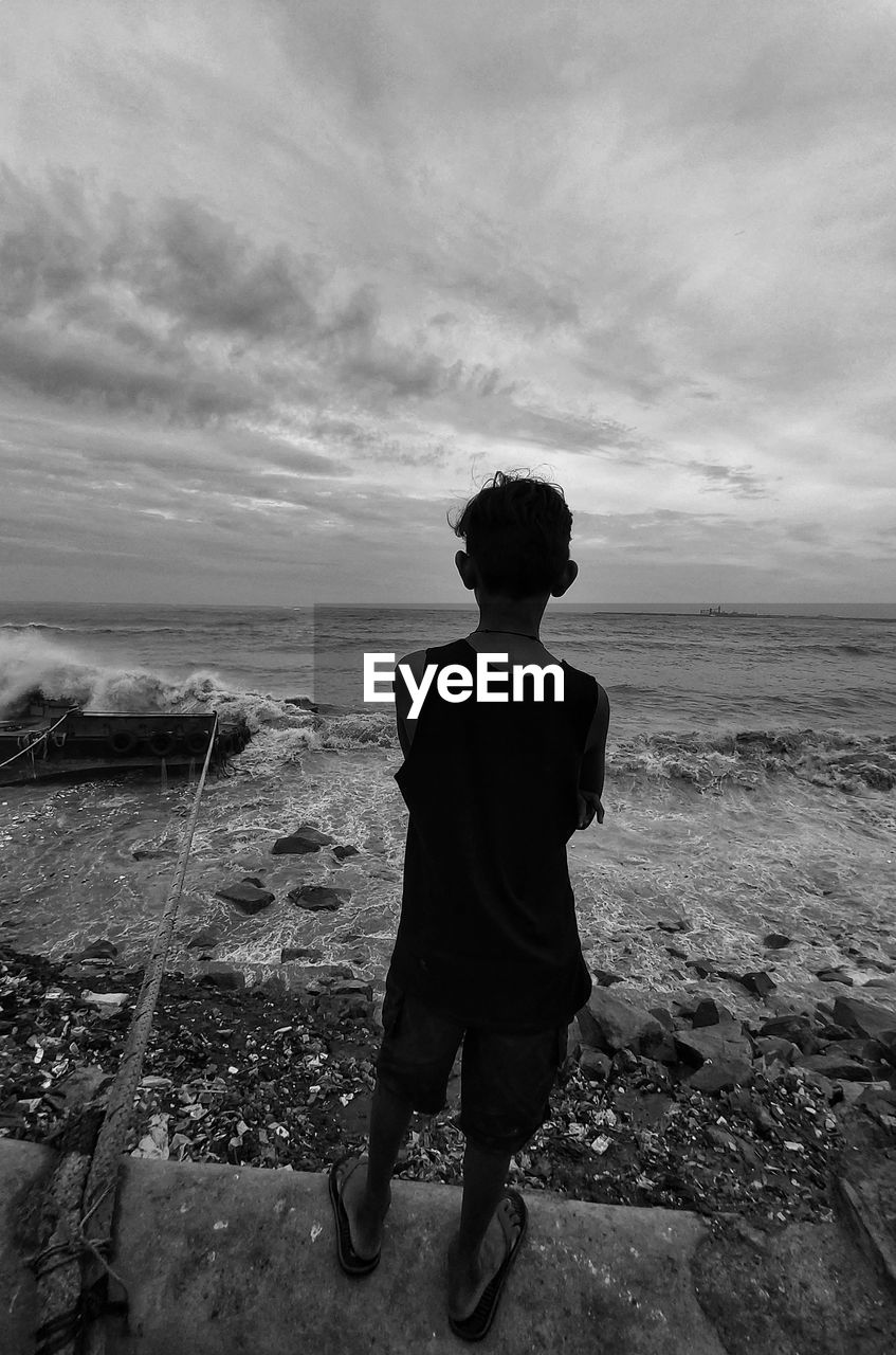 BOY STANDING ON BEACH AGAINST SKY