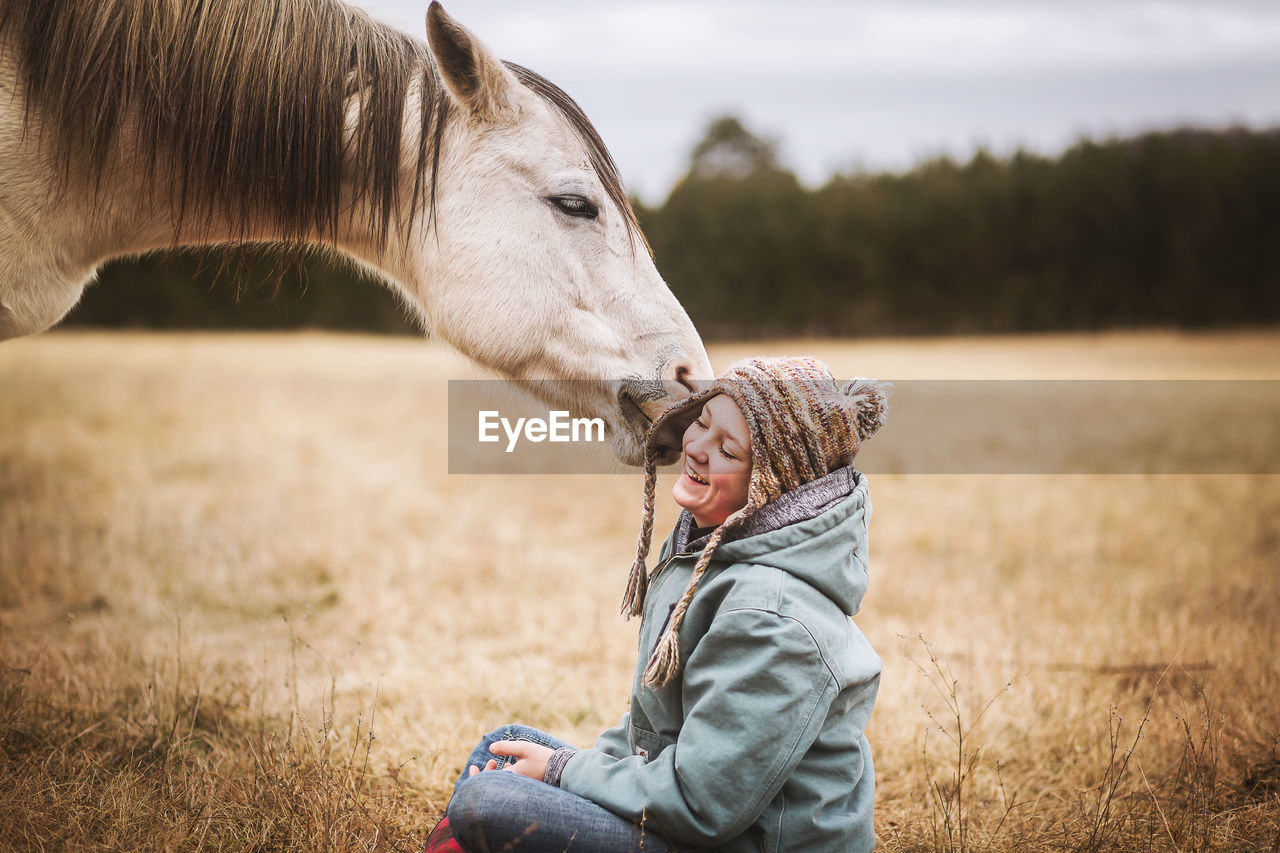 White horse playing with young girls hat while sitting in field