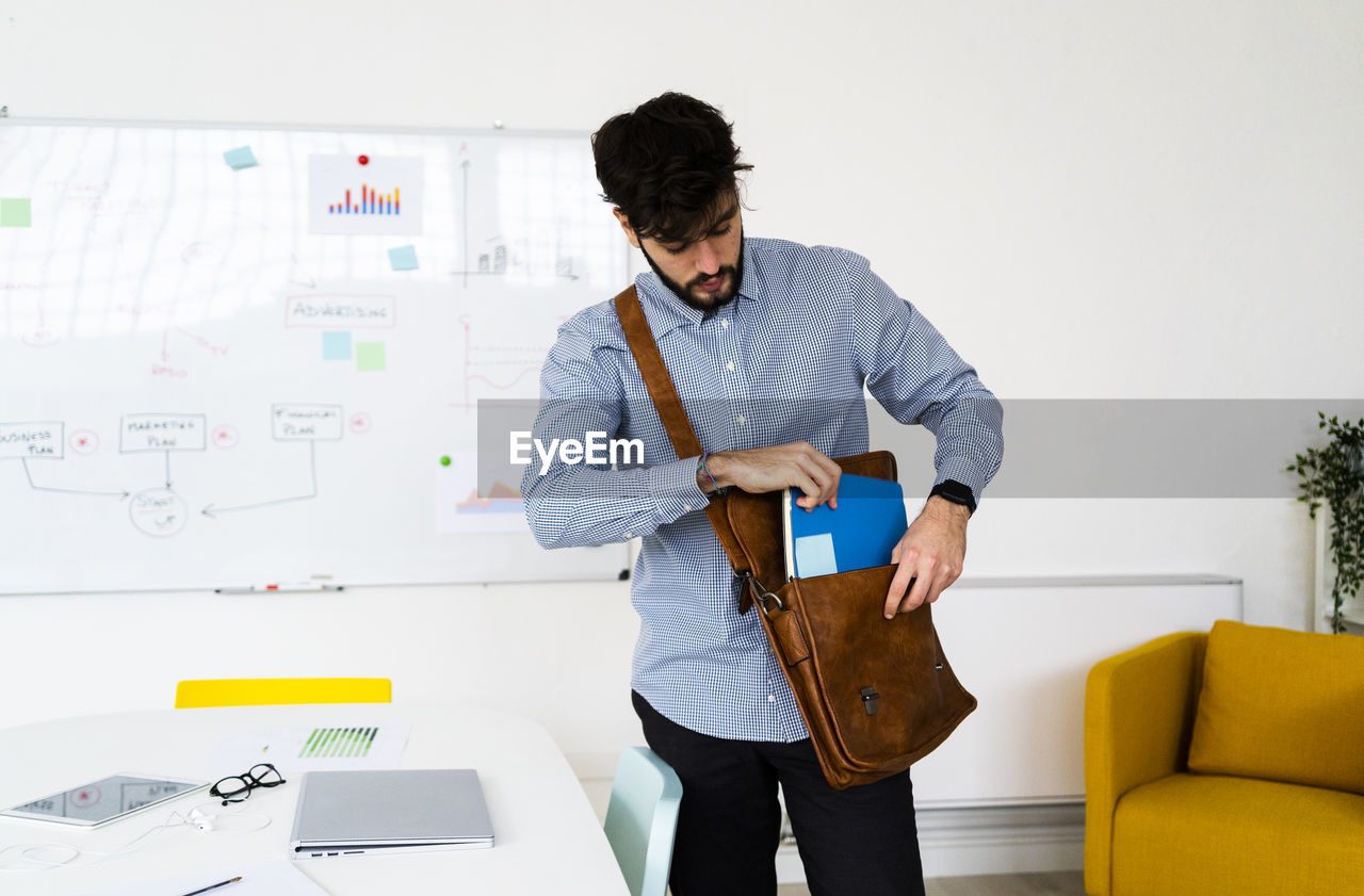 Young businessman putting notepad into shoulder bag in creative office