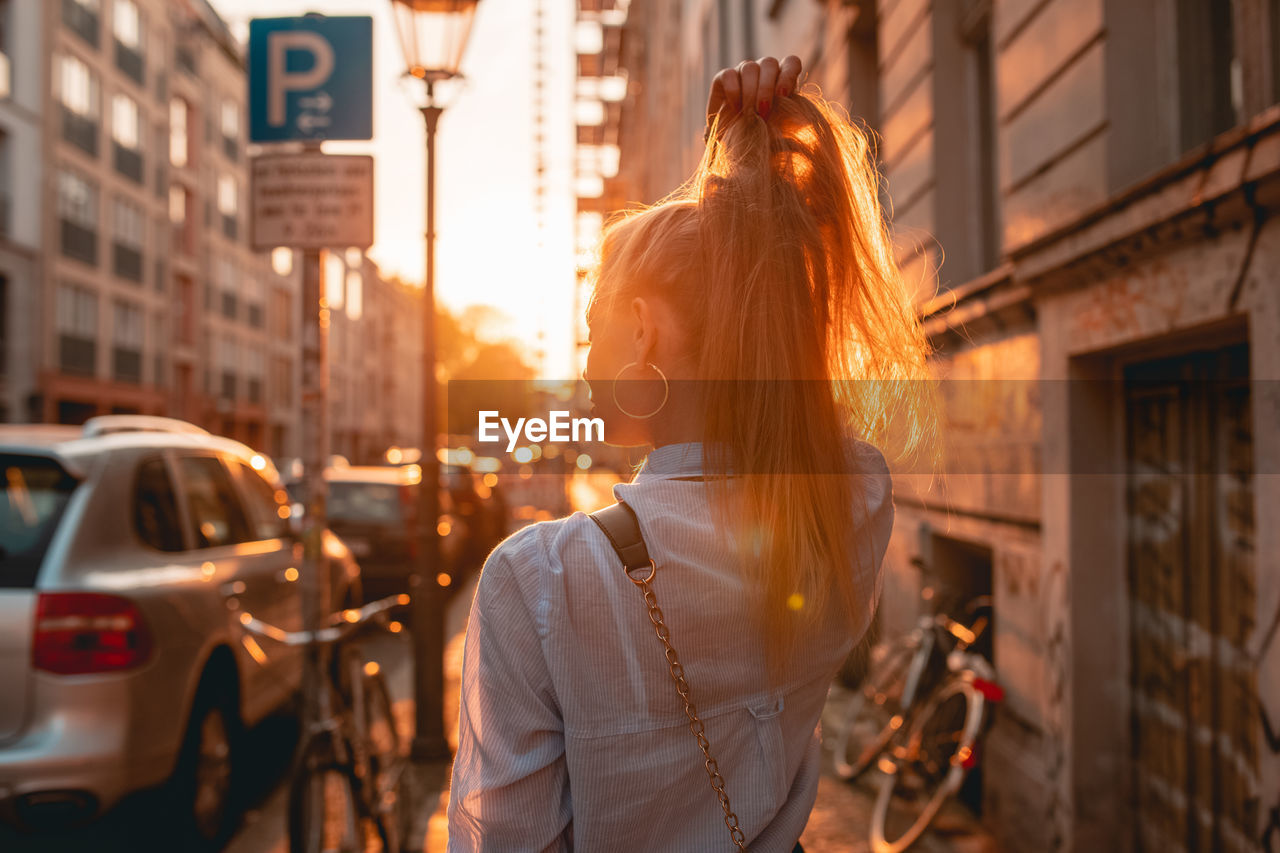 Rear view of young woman with hand in hair while standing on sidewalk during sunset