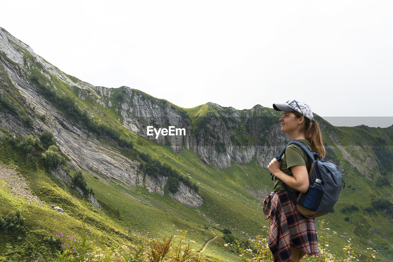 Young woman hiker in cap with backpack looking at mountain view in summer healthy active lifestyle