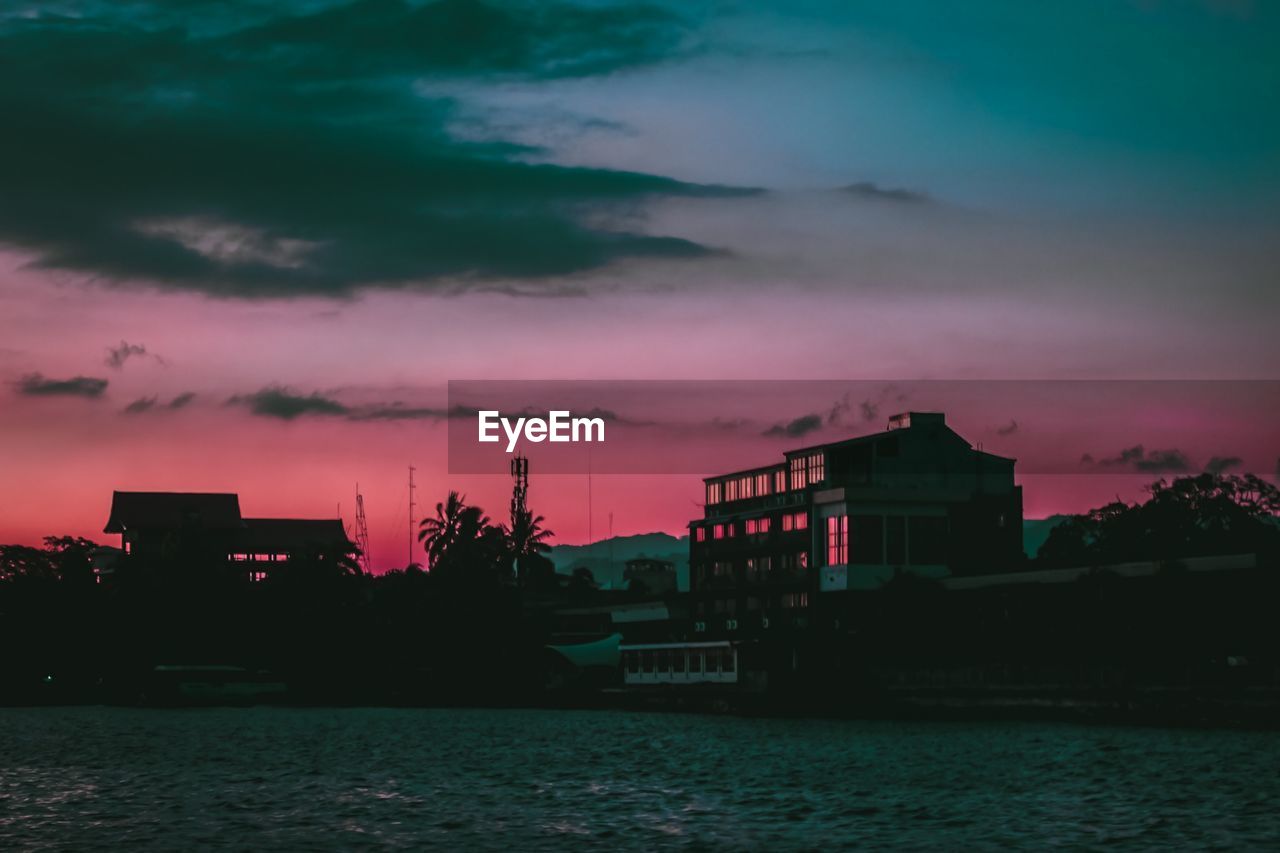 Silhouette buildings by sea against dramatic sky