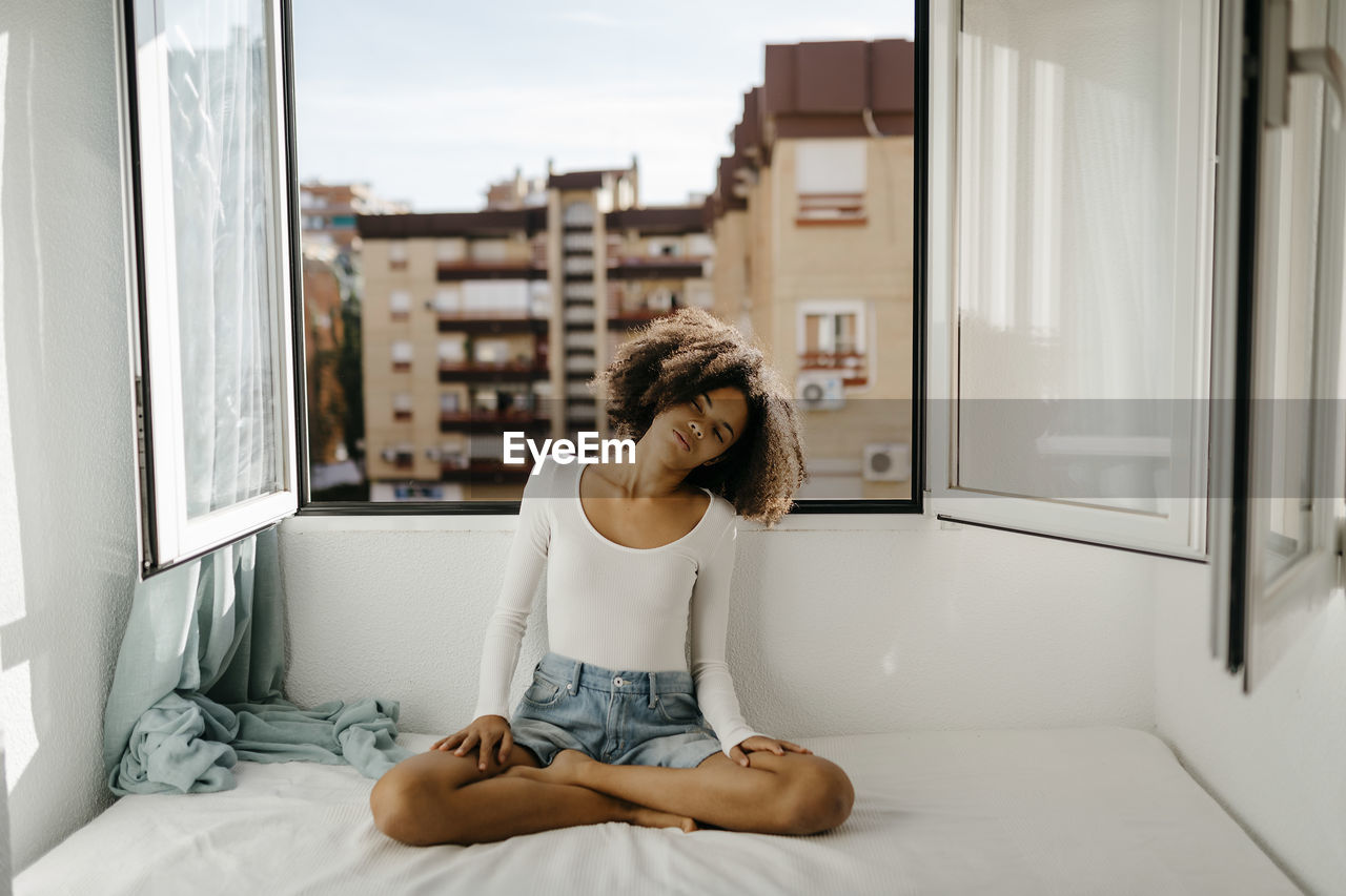 Curly haired woman sitting cross-legged on bed against window at home