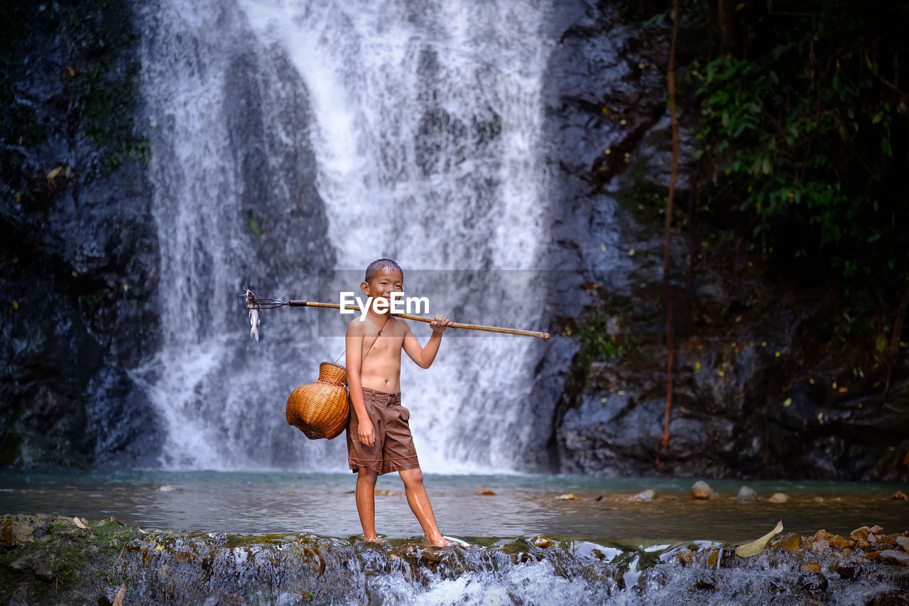 Full length of shirtless boy standing in river against waterfall