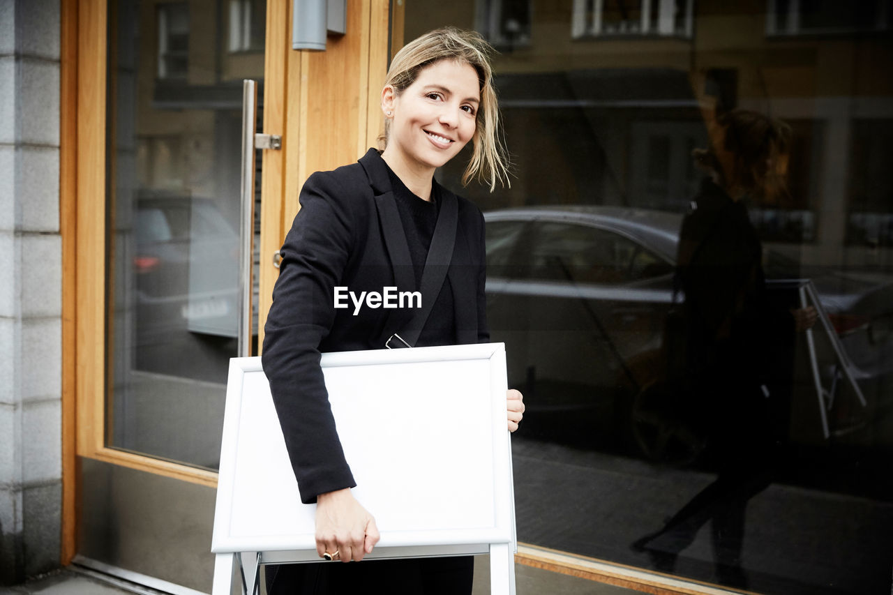 Portrait of smiling female real estate agent with blank signboard in city