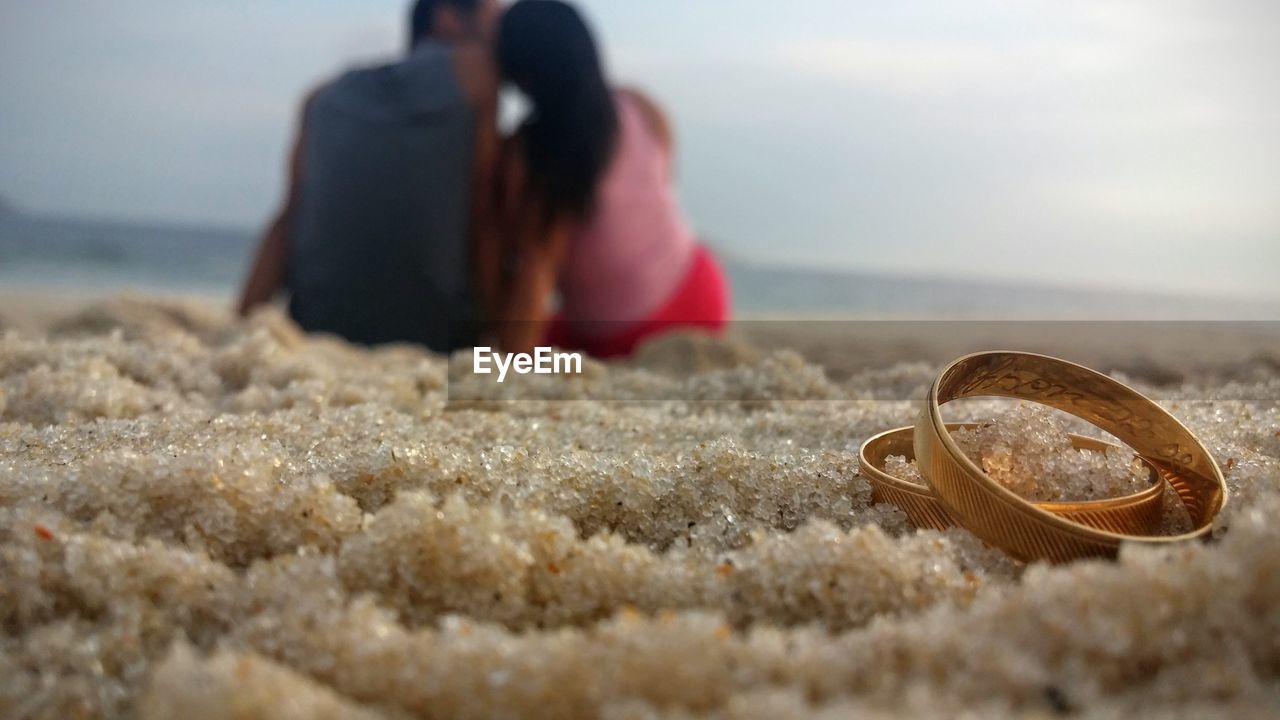 Pair of wedding rings on sand with couple sitting in background