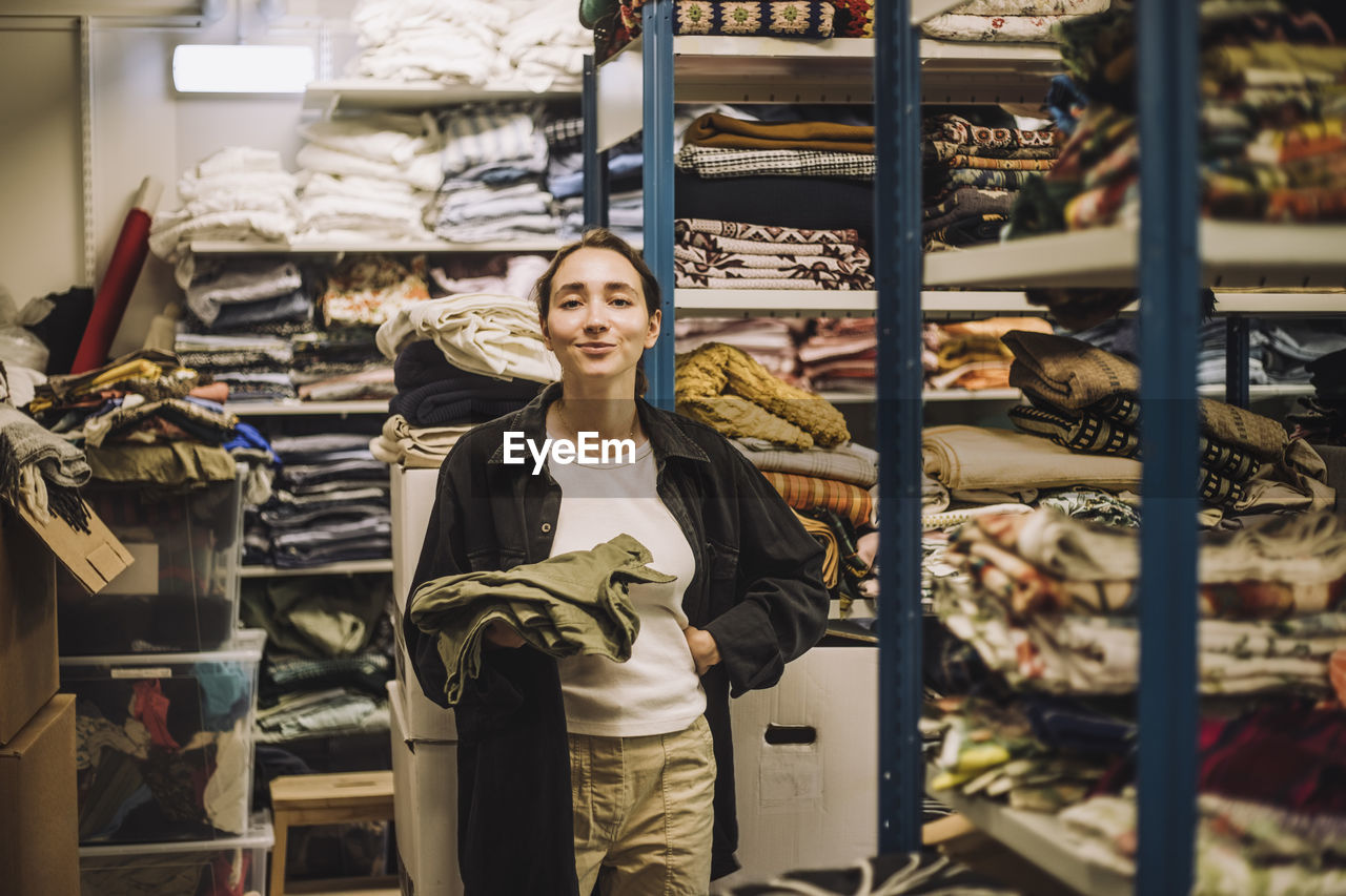 Portrait of smiling female fashion designer with hand on hip standing near clothes rack at workshop