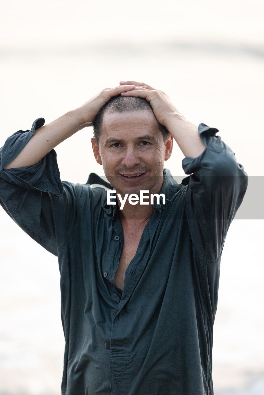 one person, adult, portrait, waist up, photo shoot, person, clothing, mature adult, front view, men, emotion, portrait photography, arm, looking at camera, arms raised, lifestyles, exercising, hand, human face, sports, smiling, day, nature, outdoors, standing, happiness, relaxation, casual clothing, wellbeing, hands behind head, spring, hand in hair, looking, focus on foreground, sitting, leisure activity, limb, fashion, vitality, human limb, beach