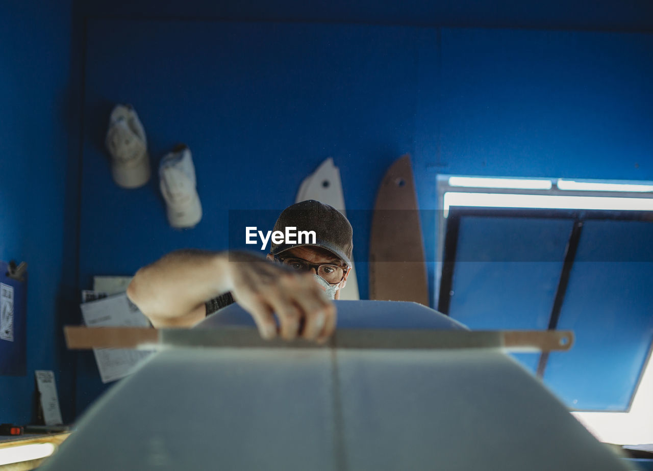 Concentrated qualified worker in protective mask aligning white surfboard in workshop with blue walls