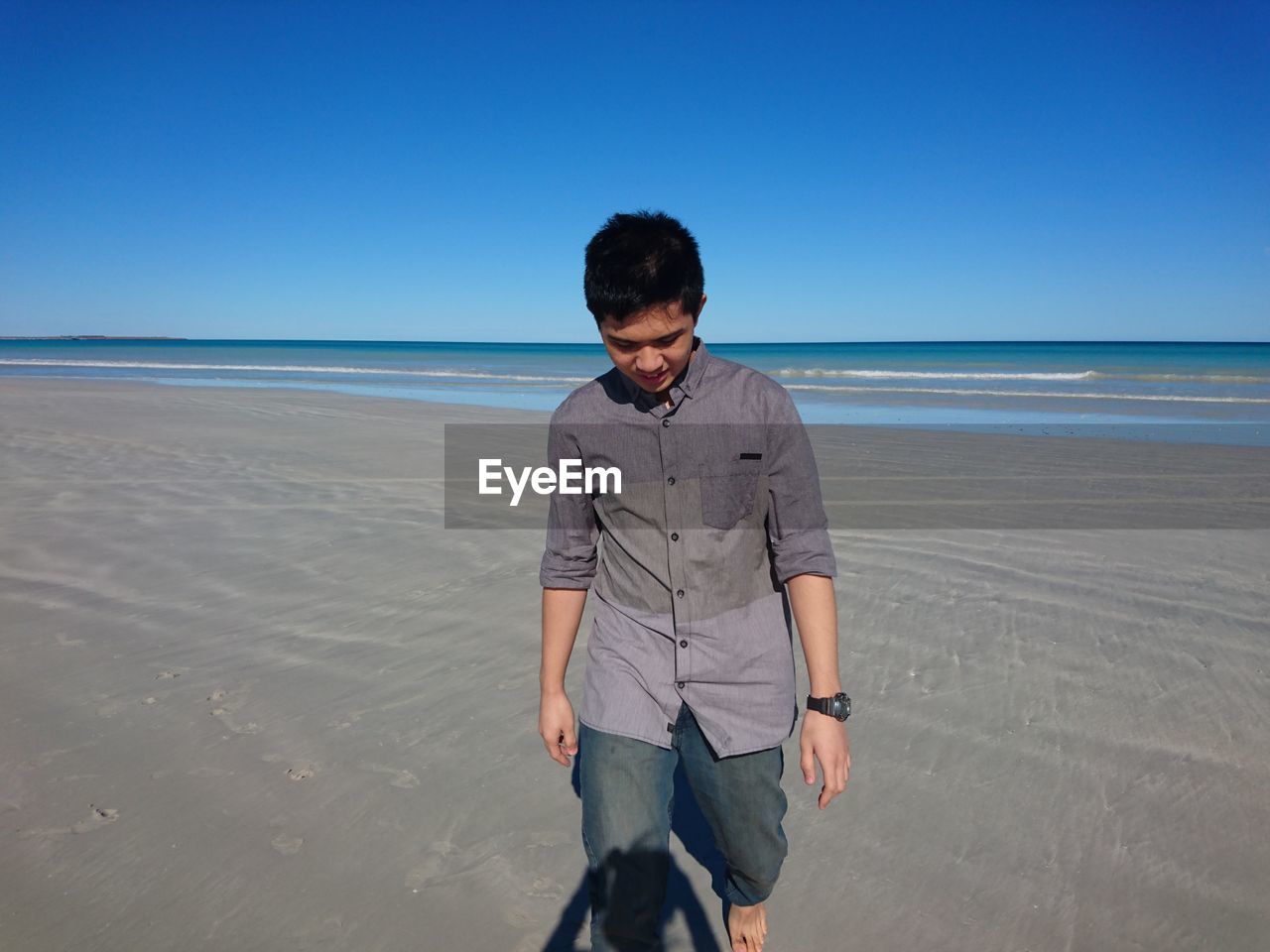 Man looking down while walking on sand at beach against clear blue sky