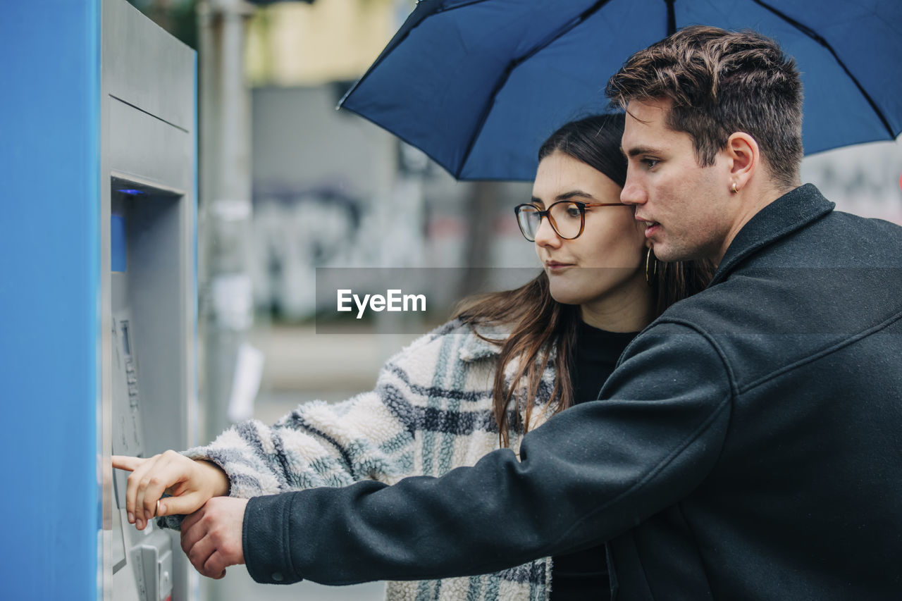 Young couple with umbrella using atm machine