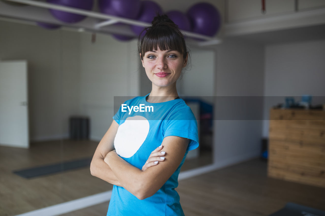 Positive young female yoga trainer in activewear standing with arms crossed and looking at camera against blurred interior of fitness studio