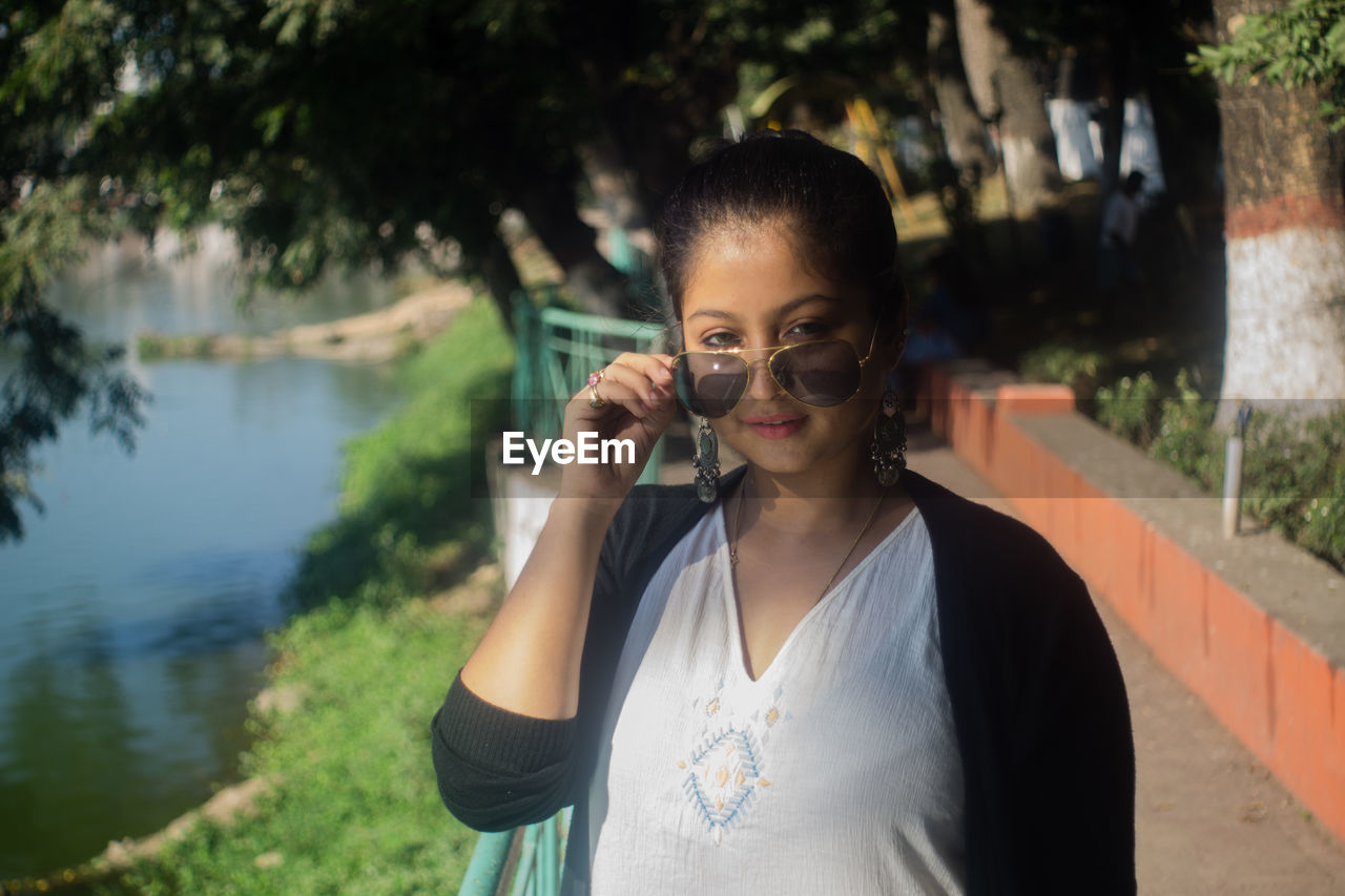 Portrait of young woman wearing sunglasses while standing against lake