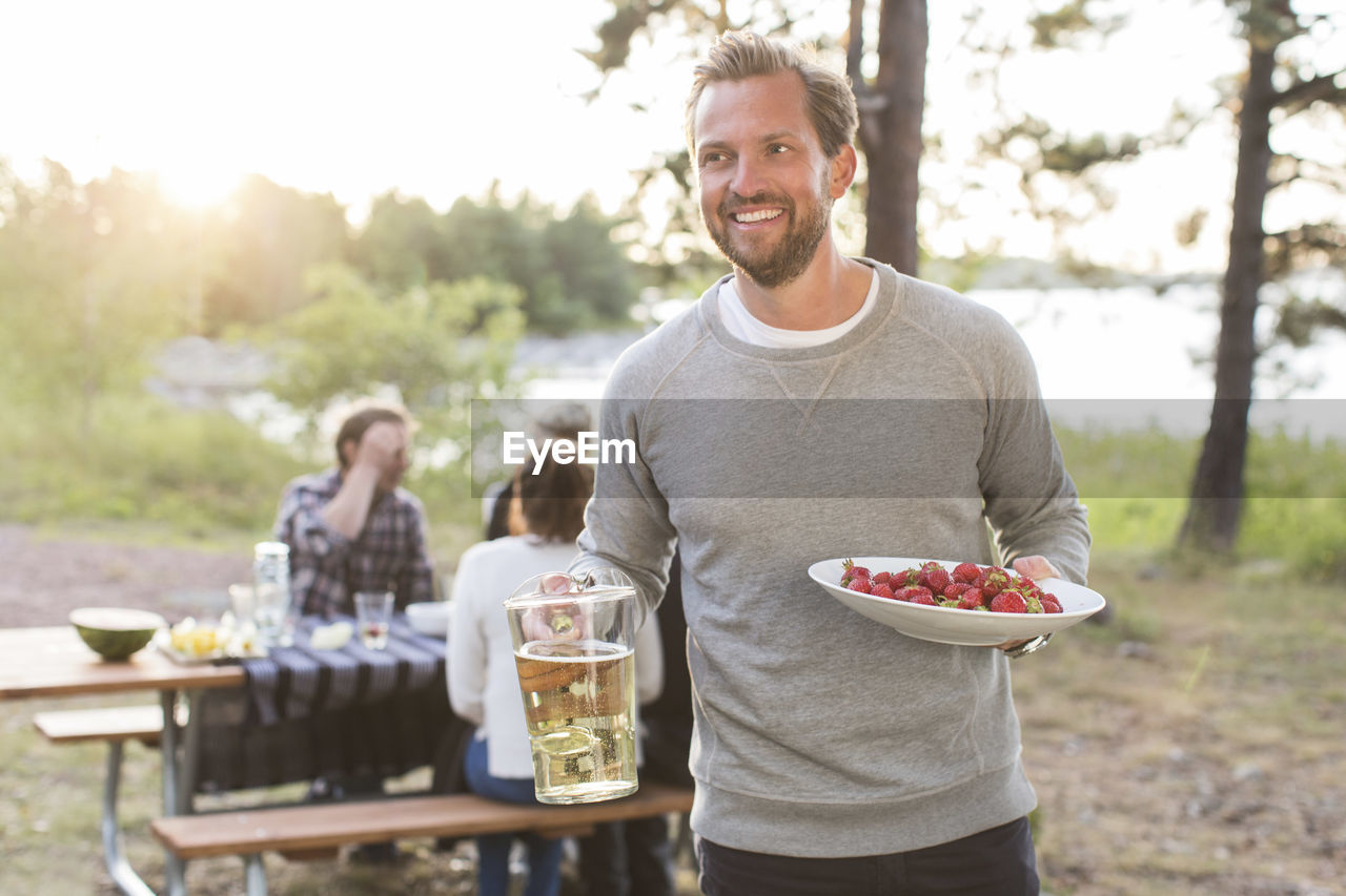 Happy man holding beer jug and strawberries with friends sitting at picnic table in background