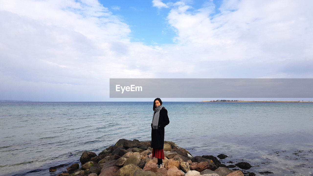 Portrait of woman standing on rocks at beach against cloudy sky