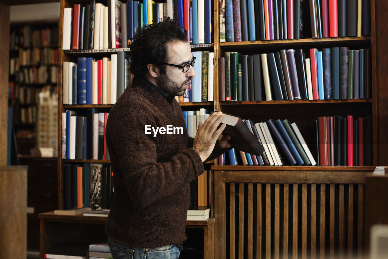 Side view of man cleaning book while standing against shelves