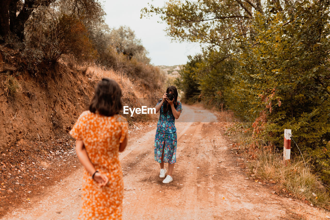 Woman photographing girlfriend while standing on dirt road in forest