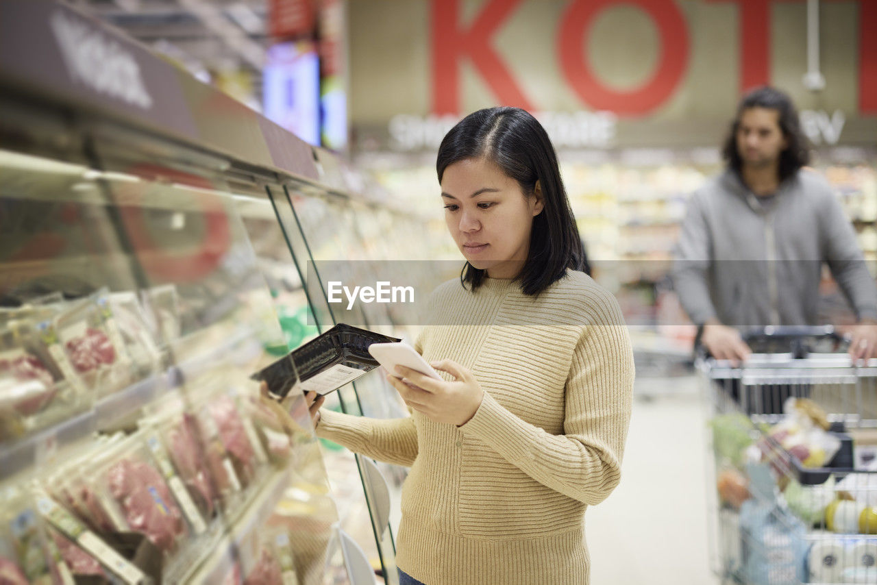 Woman doing shopping in supermarket and using cell phone to compare prices or checking shopping list