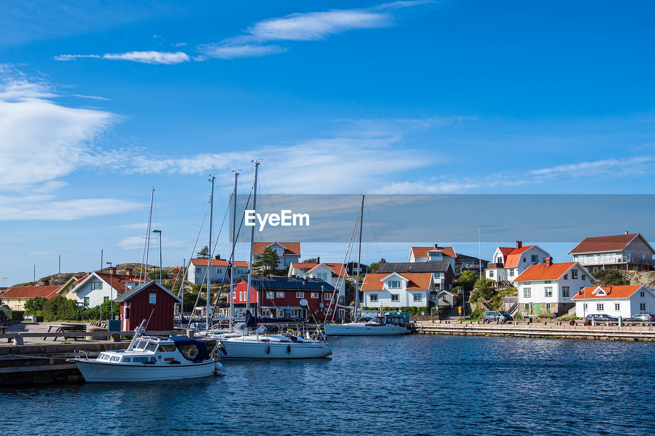 SAILBOATS MOORED IN HARBOR BY HOUSES AGAINST SKY