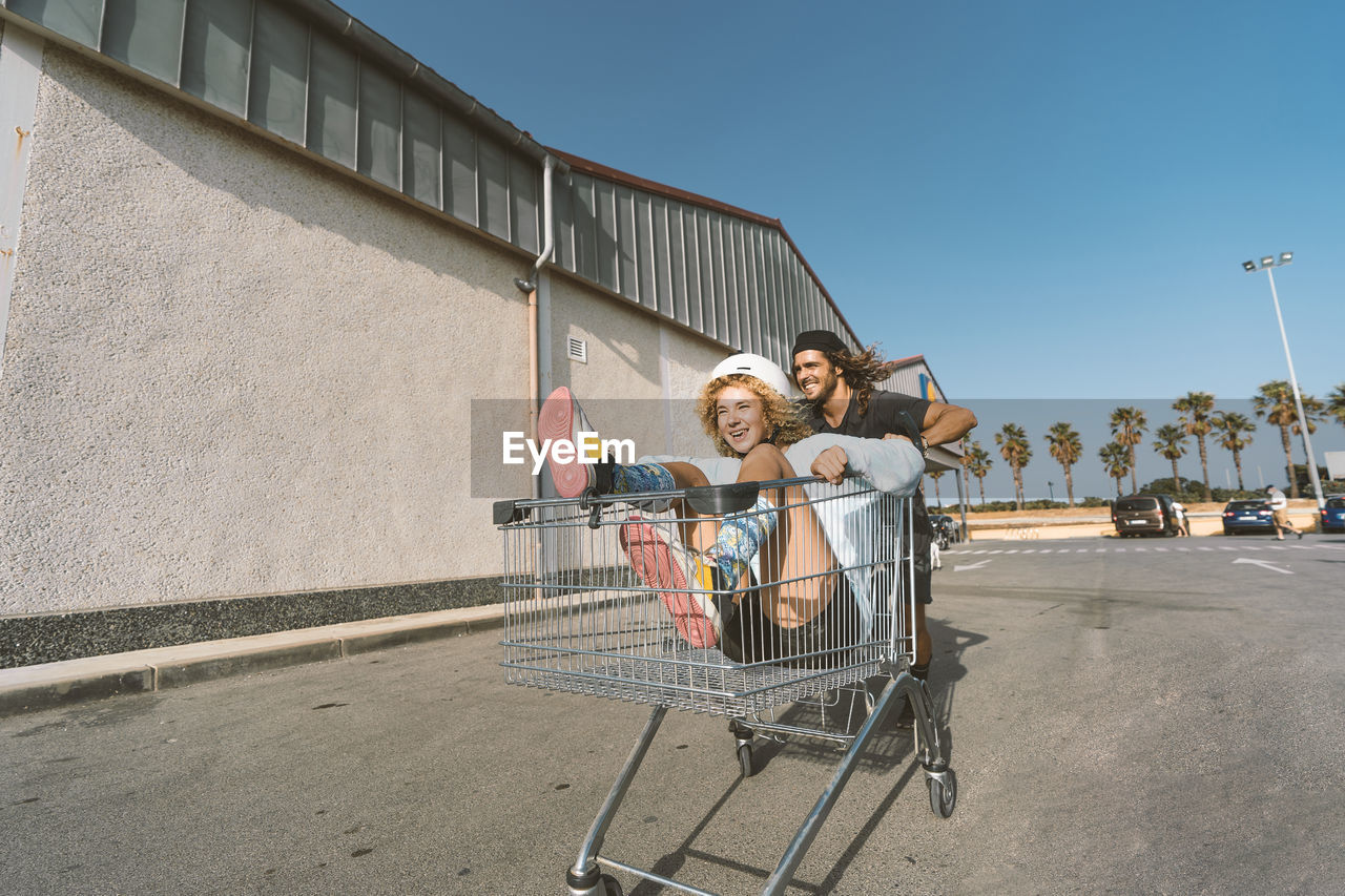 Young man pushing girlfriend sitting in shopping cart outside supermarket