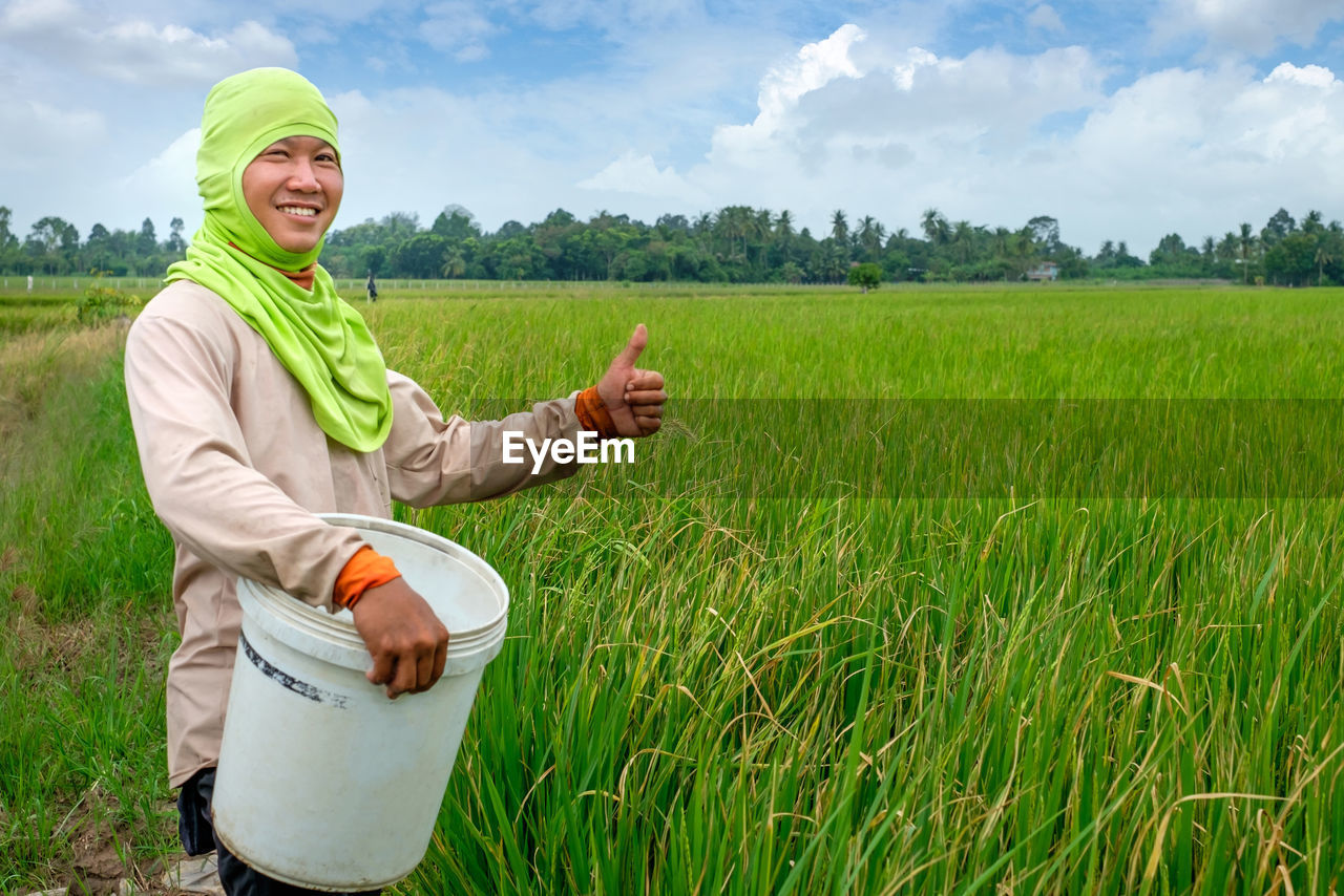 Portrait of smiling farmer gesturing thumbs up while holding bucket on agricultural field