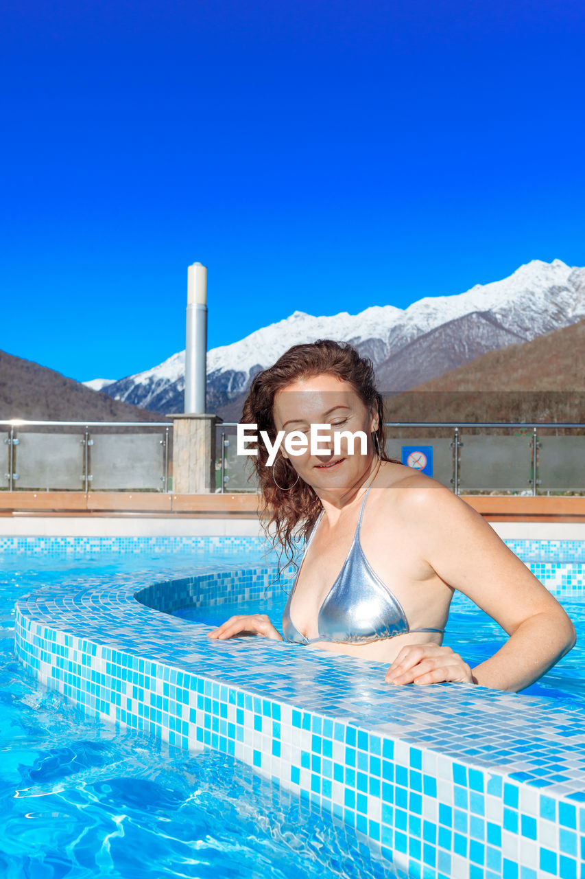 Slender woman in a silver bikini in winter by outdoor pool against the mountains and clear blue sky