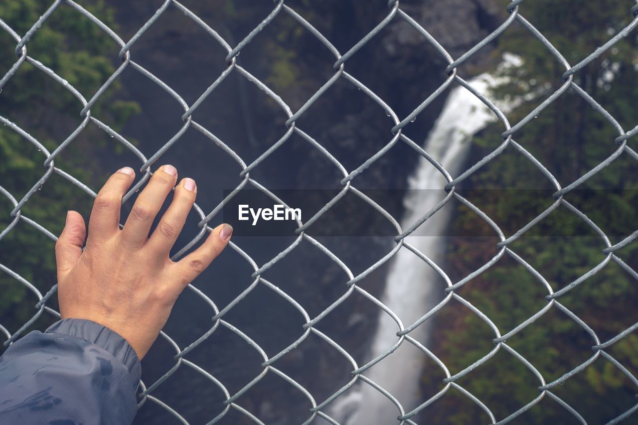 Cropped hand of person on fence against waterfall