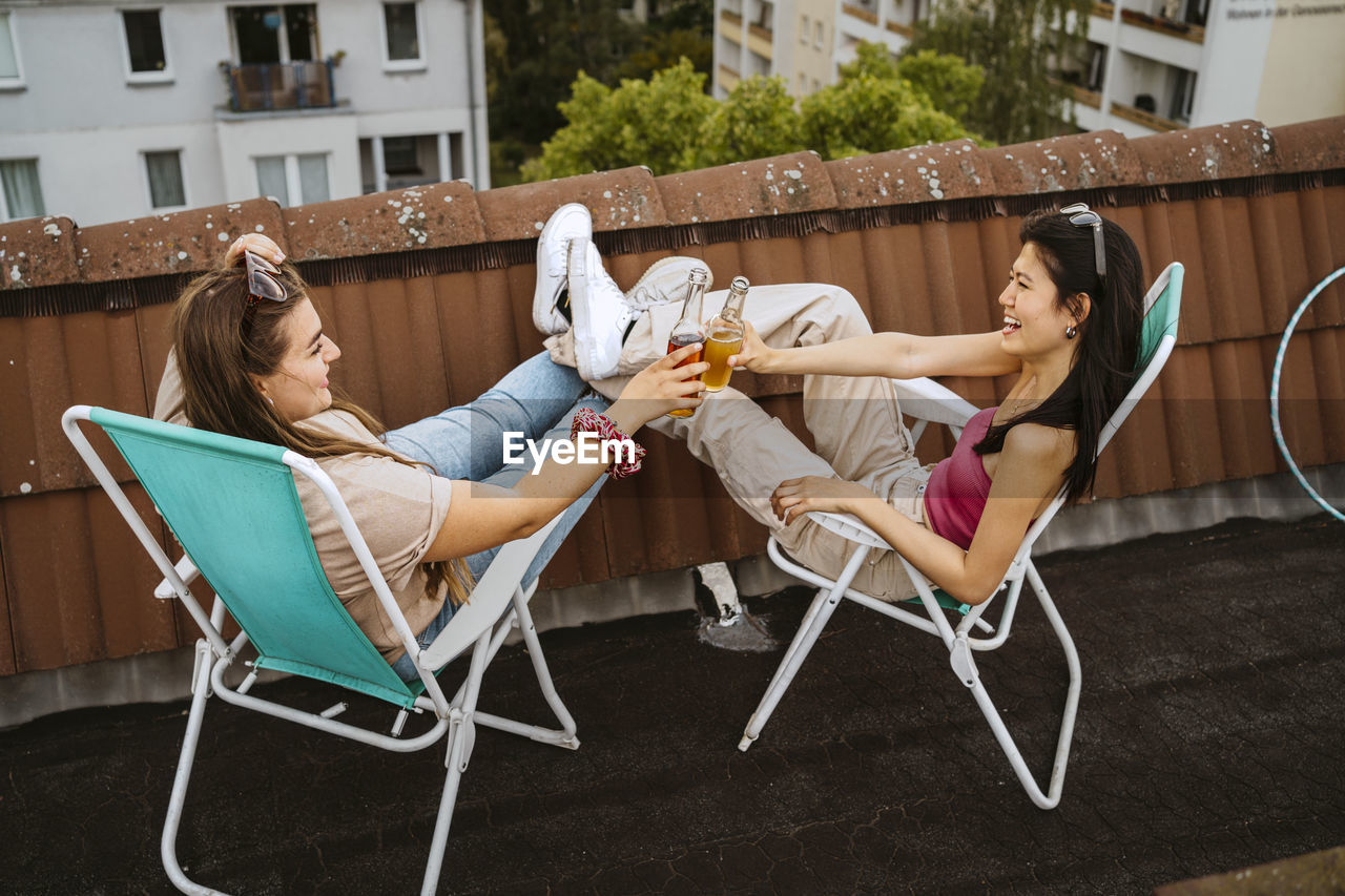 Full length of cheerful female friends toasting beer bottle while sitting on chair at rooftop