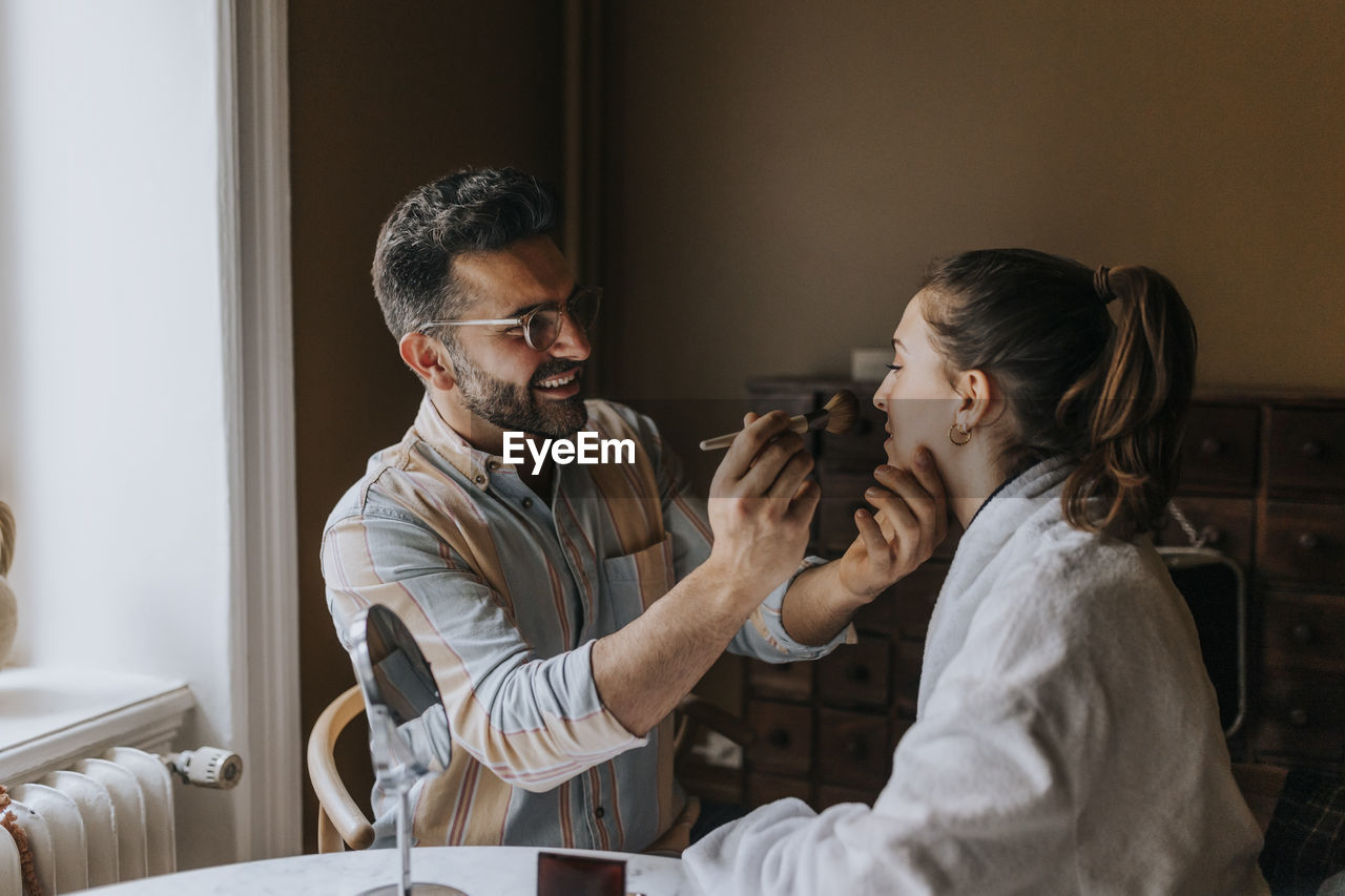 Smiling boyfriend applying make-up to non-binary person at home