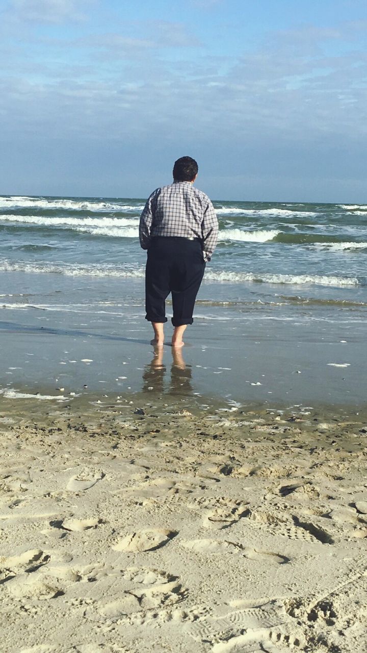 Rear view of man standing at beach against sky on sunny day
