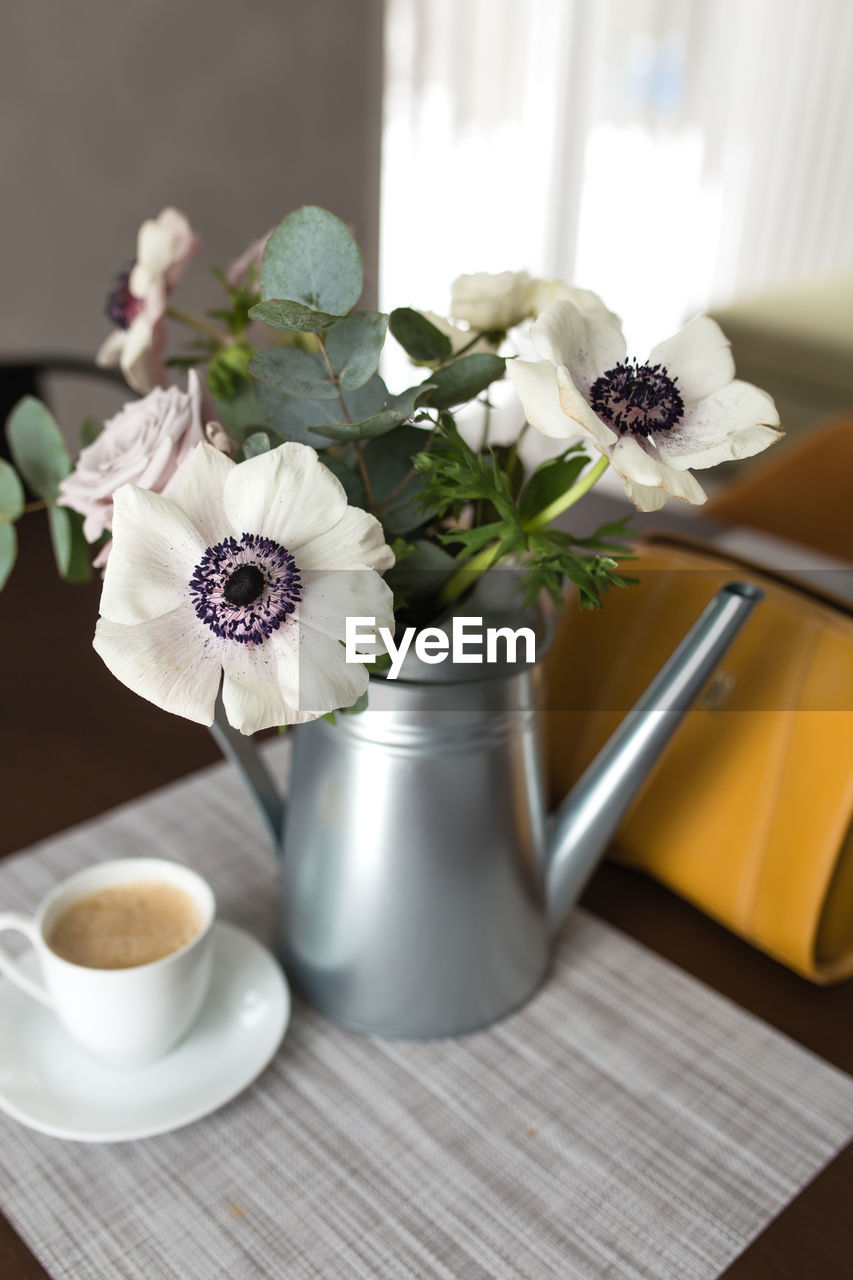 Delicate anemones flowers in a metal pot vase with a cup of coffee on the wooden table, hand bag