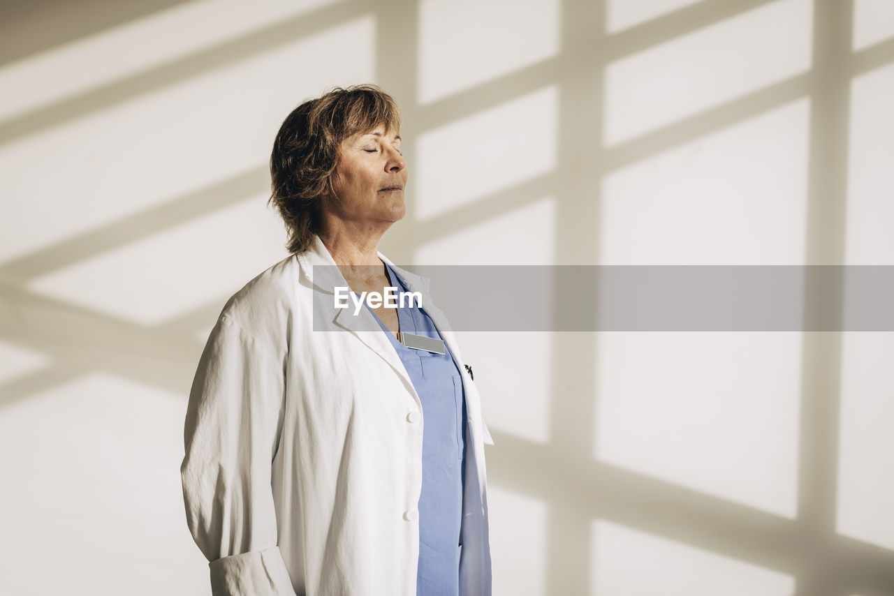 Mature female physician standing with eyes closed against wall with shadow