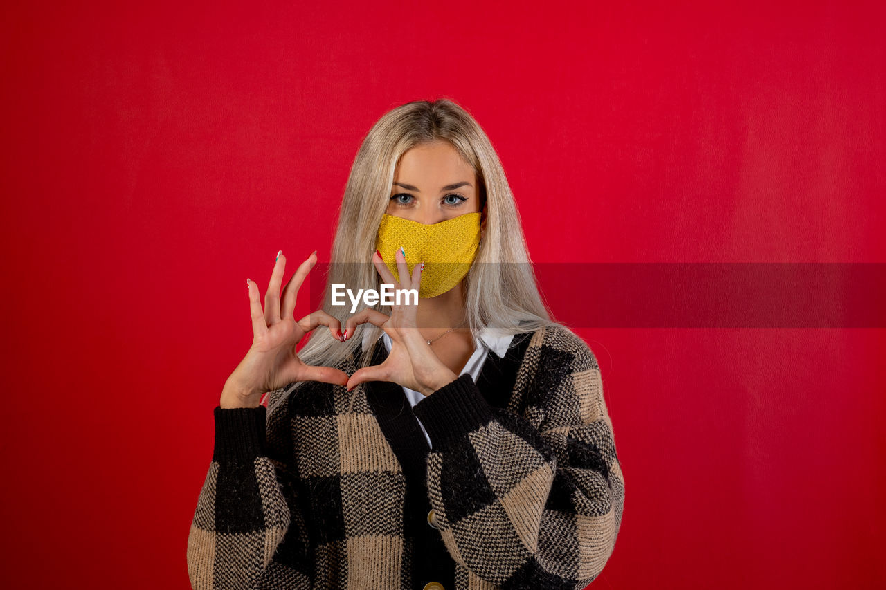 Portrait of young woman wearing mask making heart shape against red background