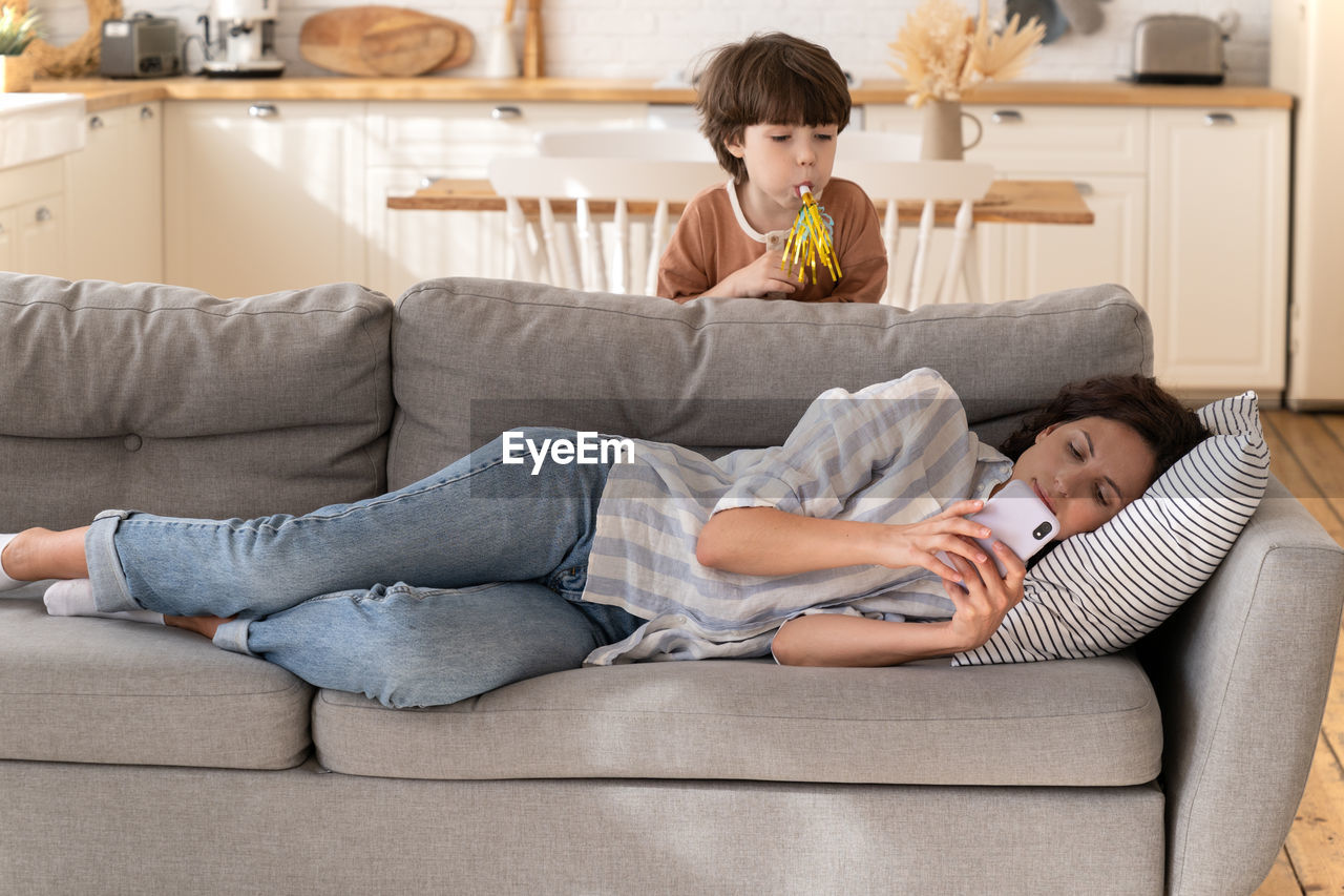 Calm young woman mother, nanny ignore noisy child lying on sofa using smartphone chatting in apps
