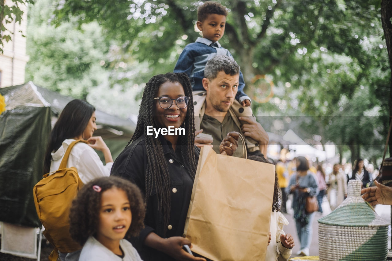 Portrait of smiling woman holding paper bag while doing shopping with family at flea market