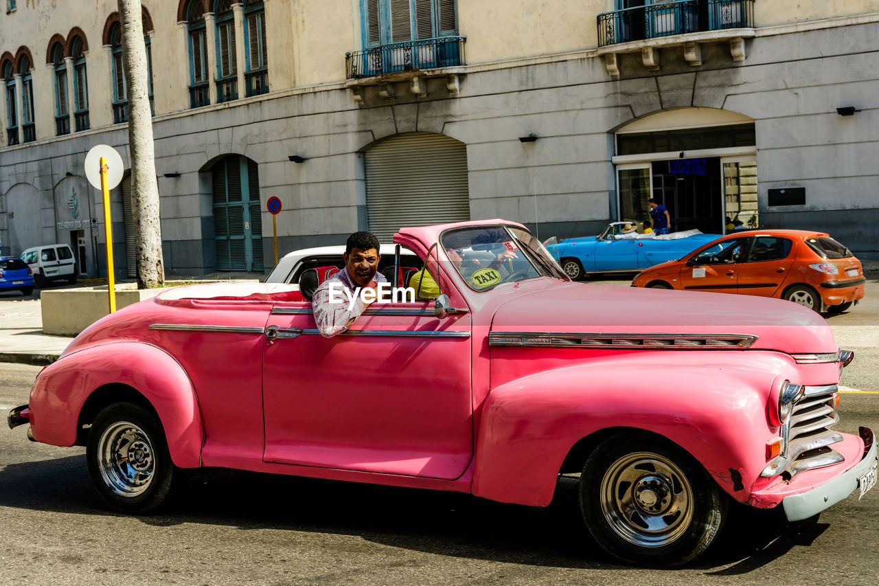 car, vehicle, mode of transportation, transportation, land vehicle, motor vehicle, architecture, antique car, city, building exterior, street, retro styled, built structure, vintage car, automobile, red, travel, driving, pink, old, history, day, the past, taxi, convertible, road, city street, city life, travel destinations, adult