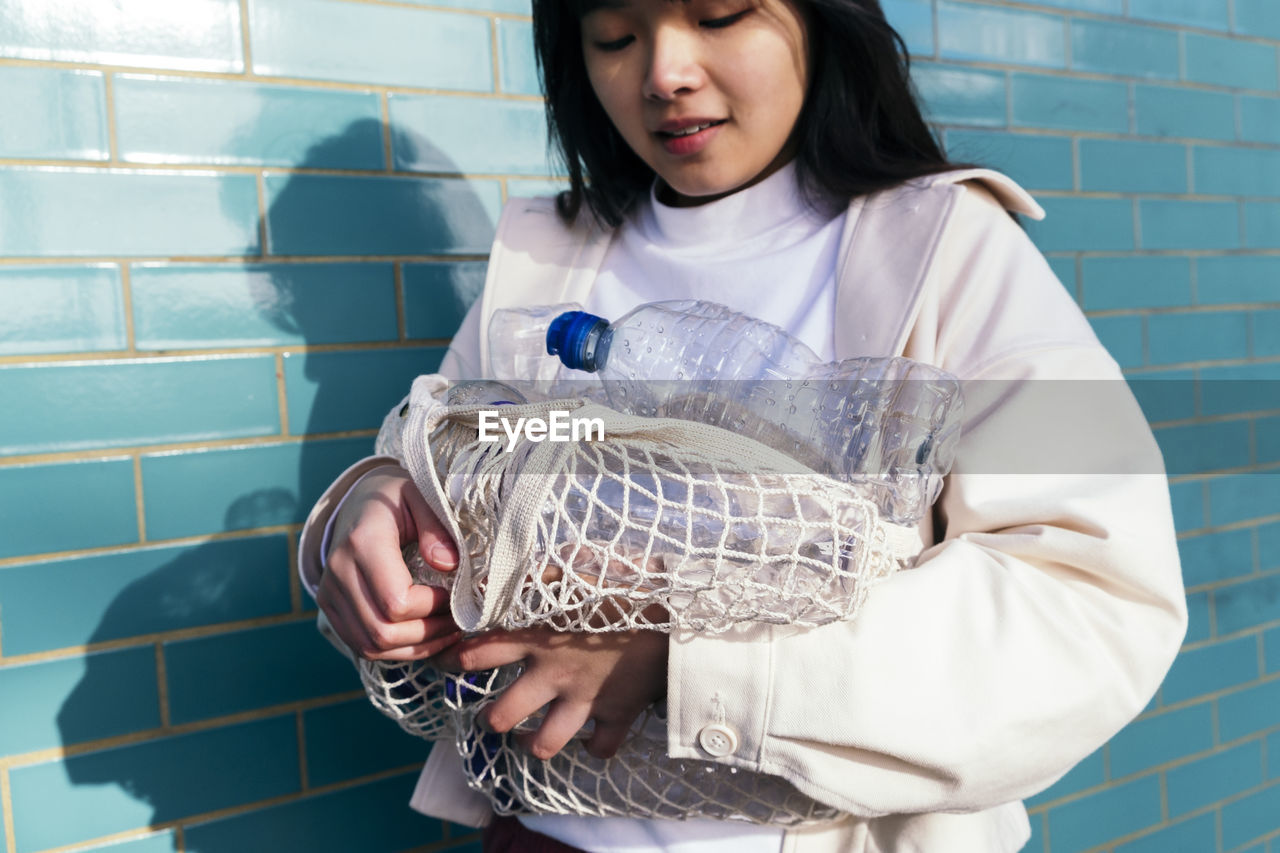 Young woman holding empty plastic bottles in mesh bag by turquoise brick wall