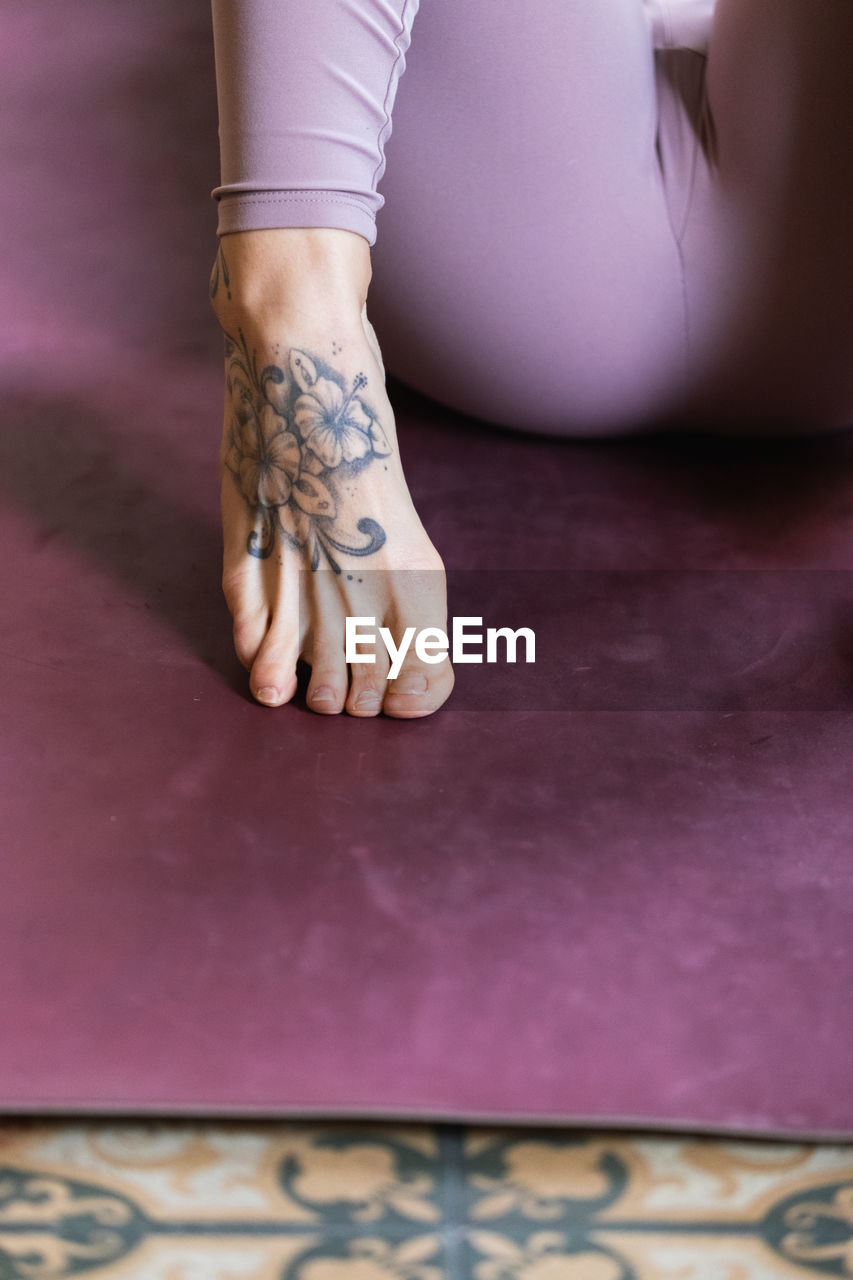 Crop anonymous barefoot of female ith tattoo on foot in leggings lying on yoga mat on the floor at home