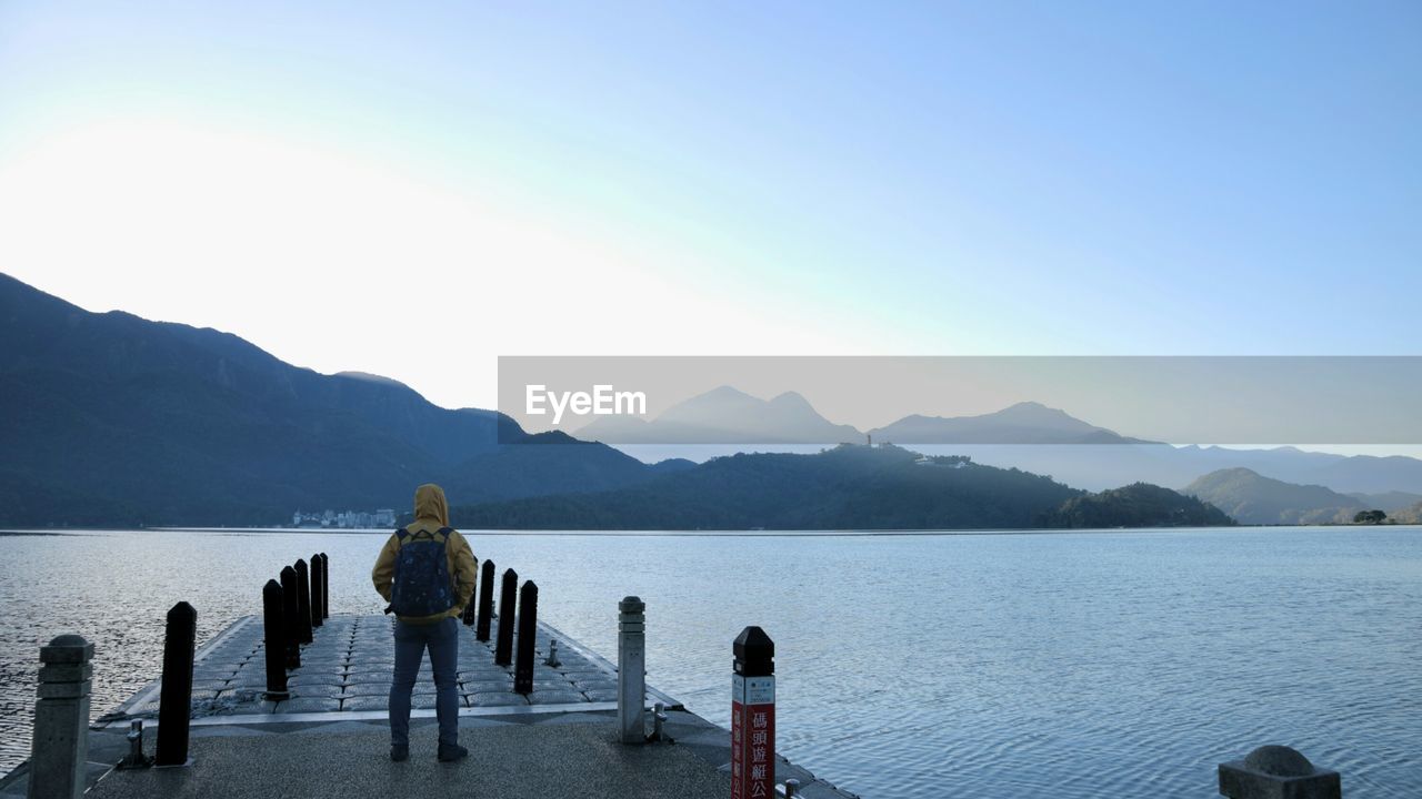 Rear view of man standing on pier over lake against clear sky