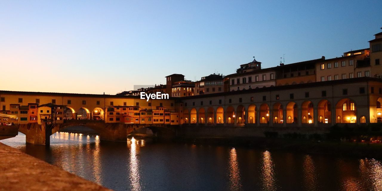 BRIDGE OVER RIVER BY ILLUMINATED BUILDINGS AGAINST SKY