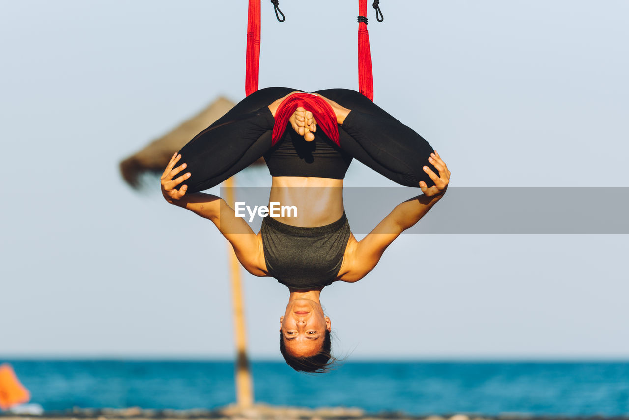 Woman practicing aerial yoga on silk at beach against sky