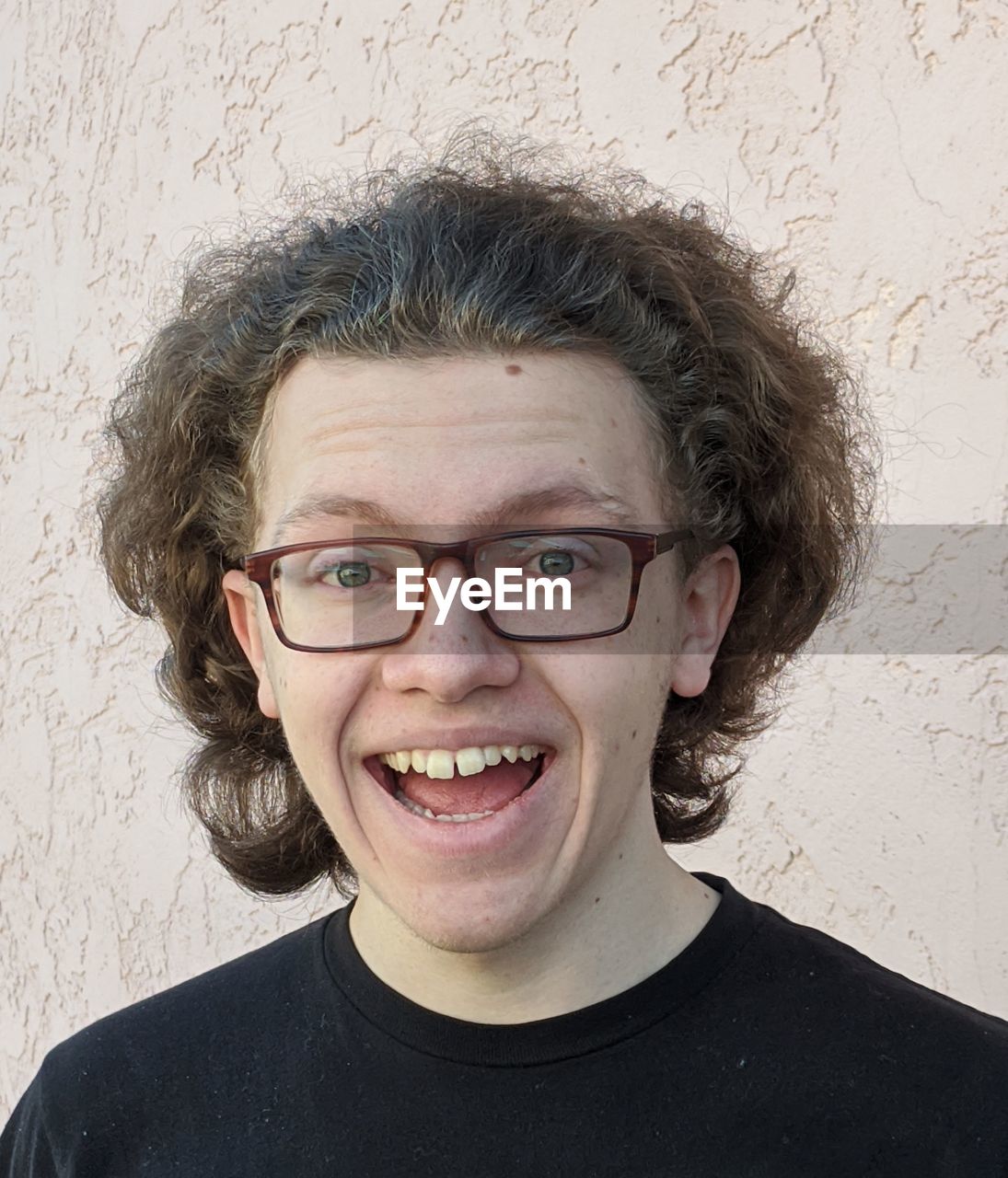 Portrait of smiling teenage boy with curly hair and glasses against wall