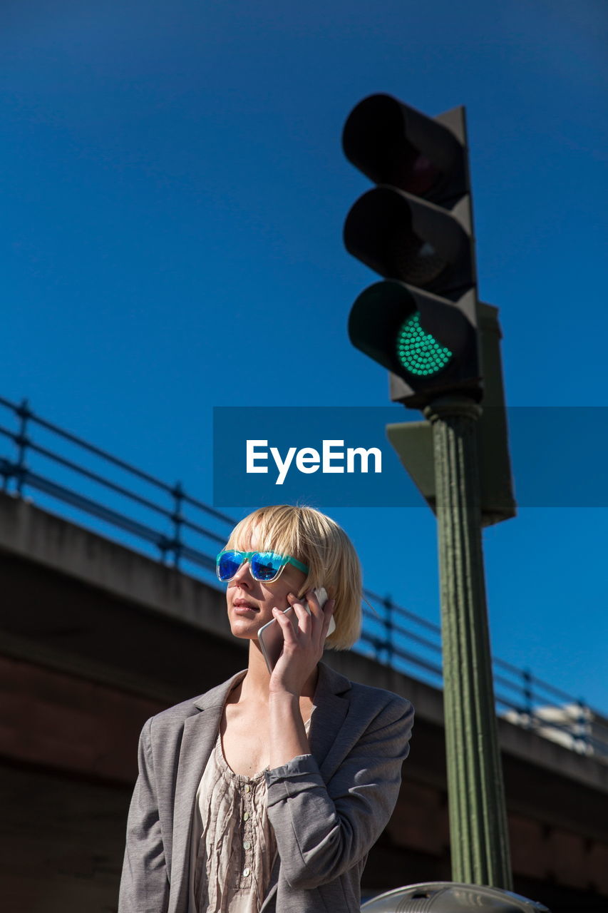 Blonde businesswoman in sunglasses talking on smartphone and looking away at green traffic light on street