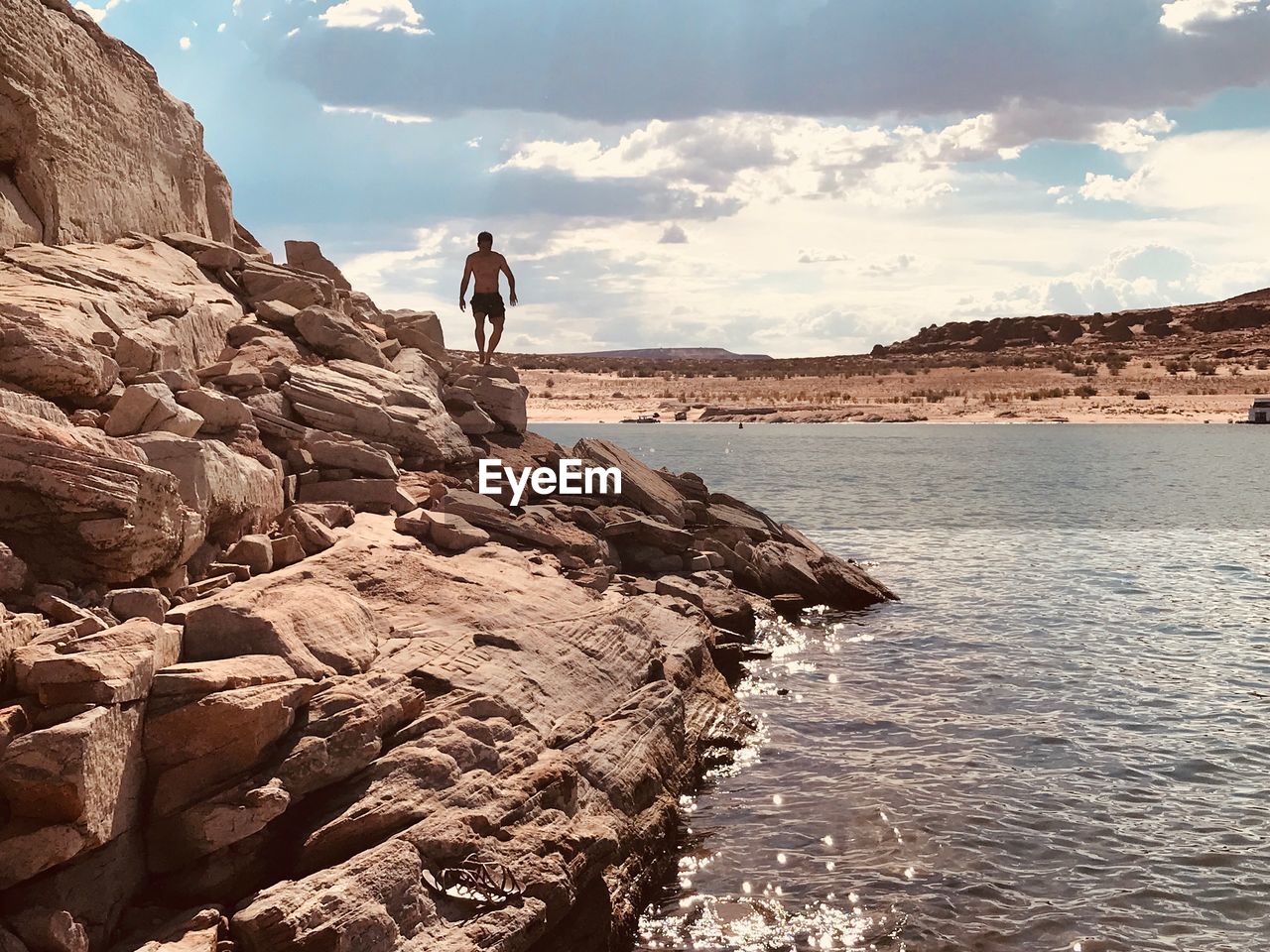 REAR VIEW OF MAN STANDING ON ROCK BY SEA AGAINST SKY