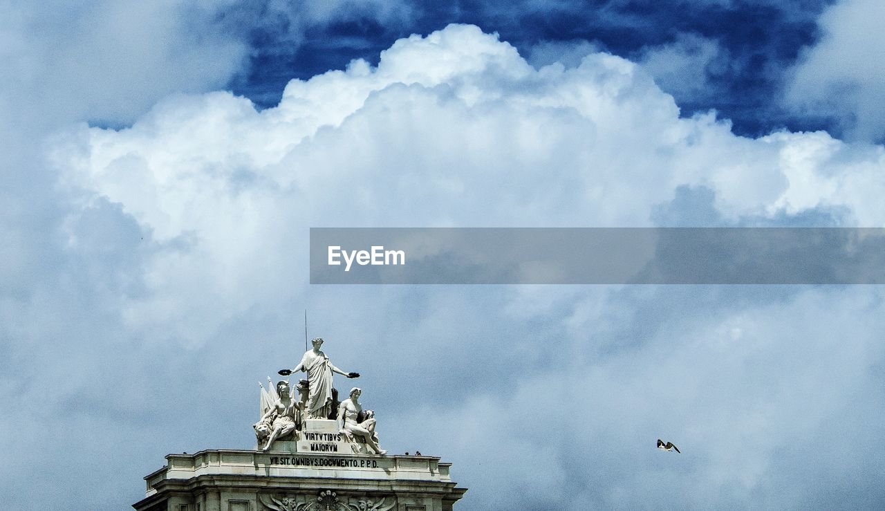 Low angle view of bird flying by statues at rua augusta arch against cloudy sky