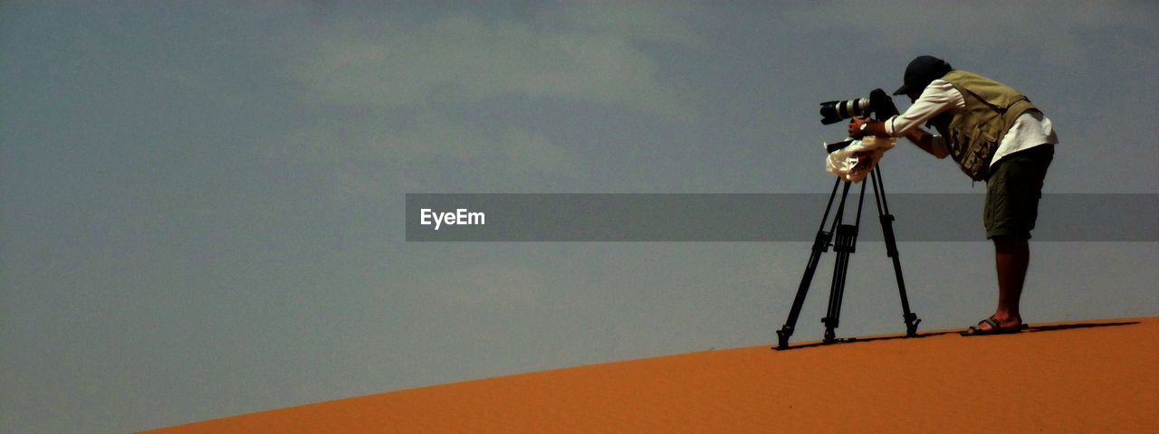 Side view full length of photographer with tripod at desert against sky