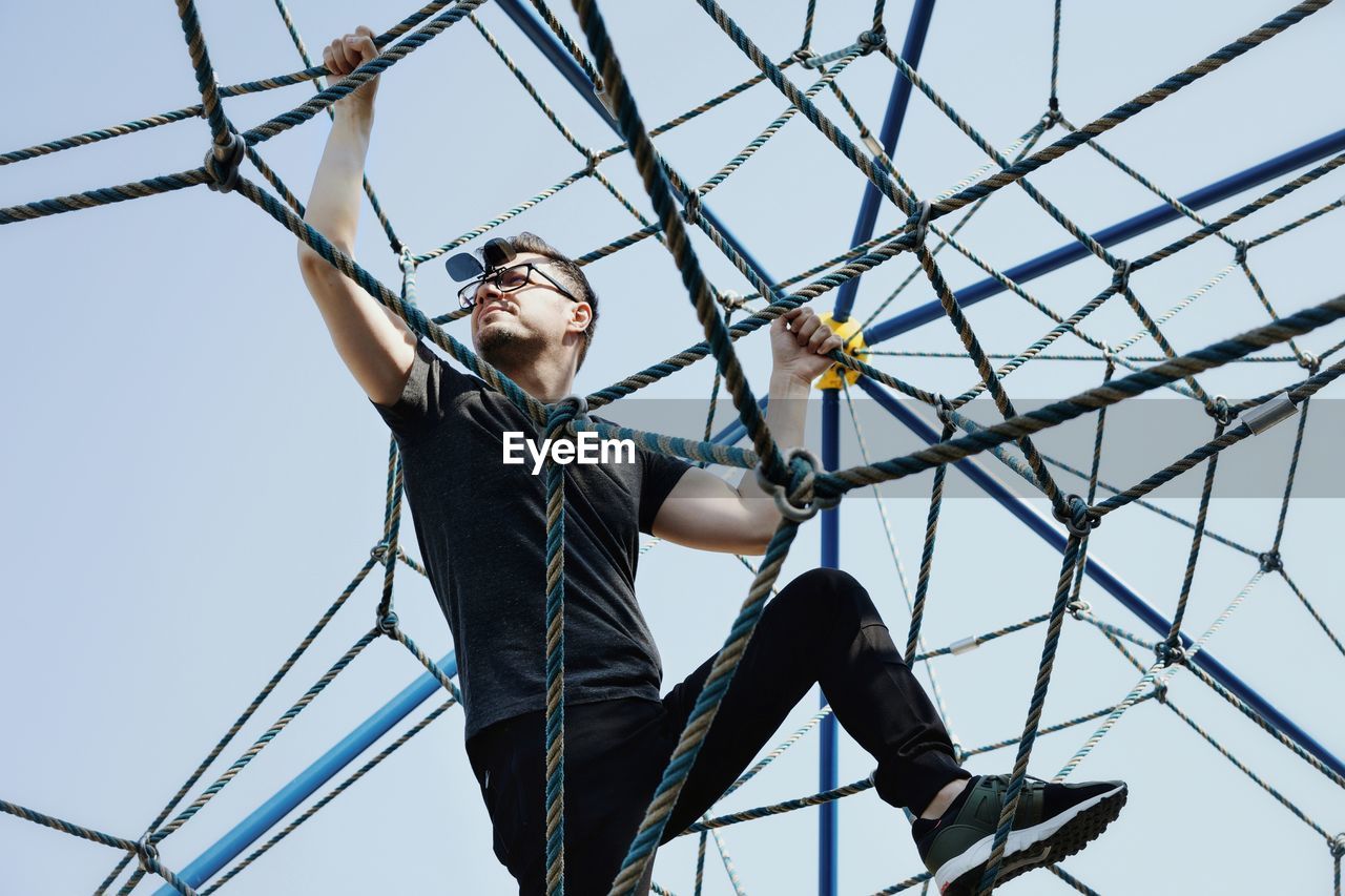 Low angle view of mid adult man climbing rope against clear sky