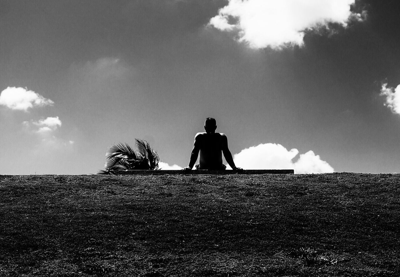 Rear view of silhouette man sitting on grassy hill against sky