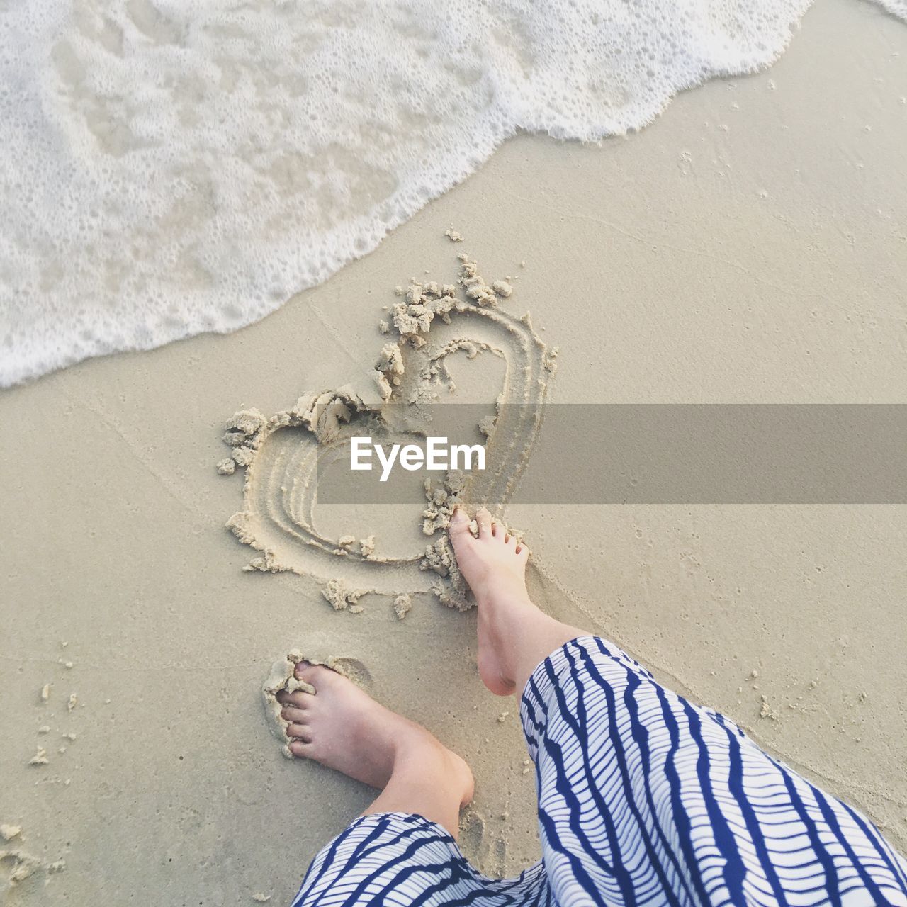 Low section of woman drawing heart shape at beach through leg